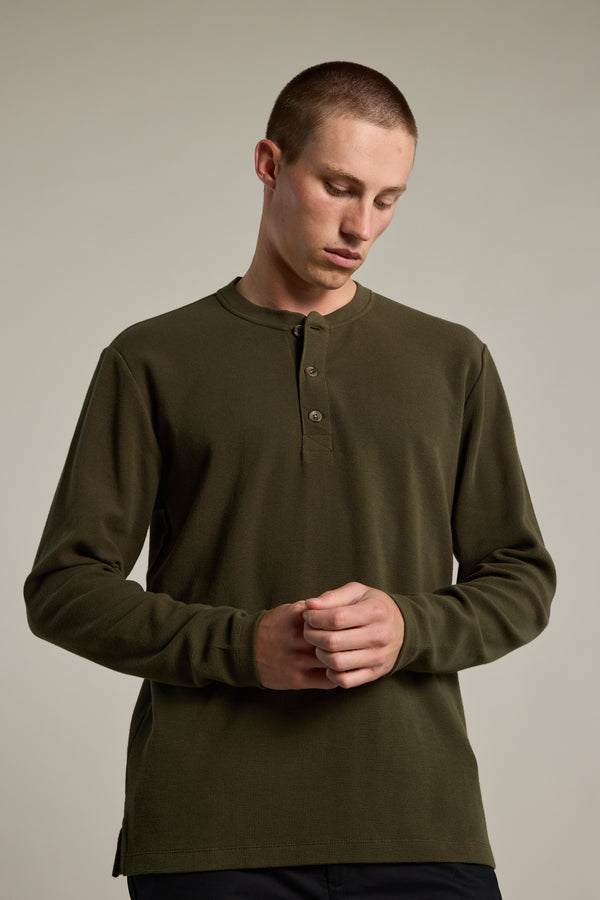 A young man with short hair models the Barkers Avenue Waffle Henley in olive green, made from organic cotton, standing against a light gray background and looking down with his hands loosely clasped in front of him.