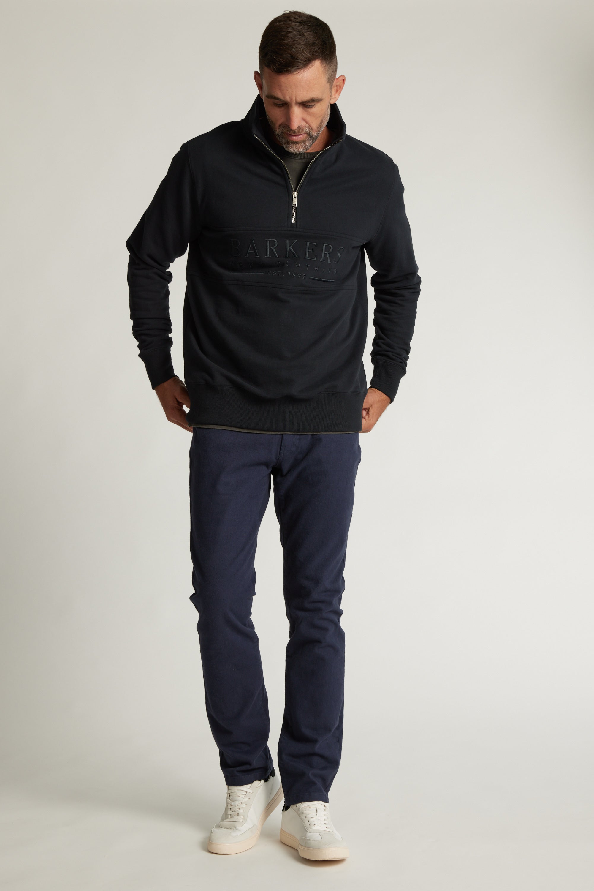 A man with short dark hair and a beard wears the Barkers Heritage 1/4 Zip sweater and dark blue pants with white sneakers. He stands against a plain light background, looking down with his hands in his pockets.