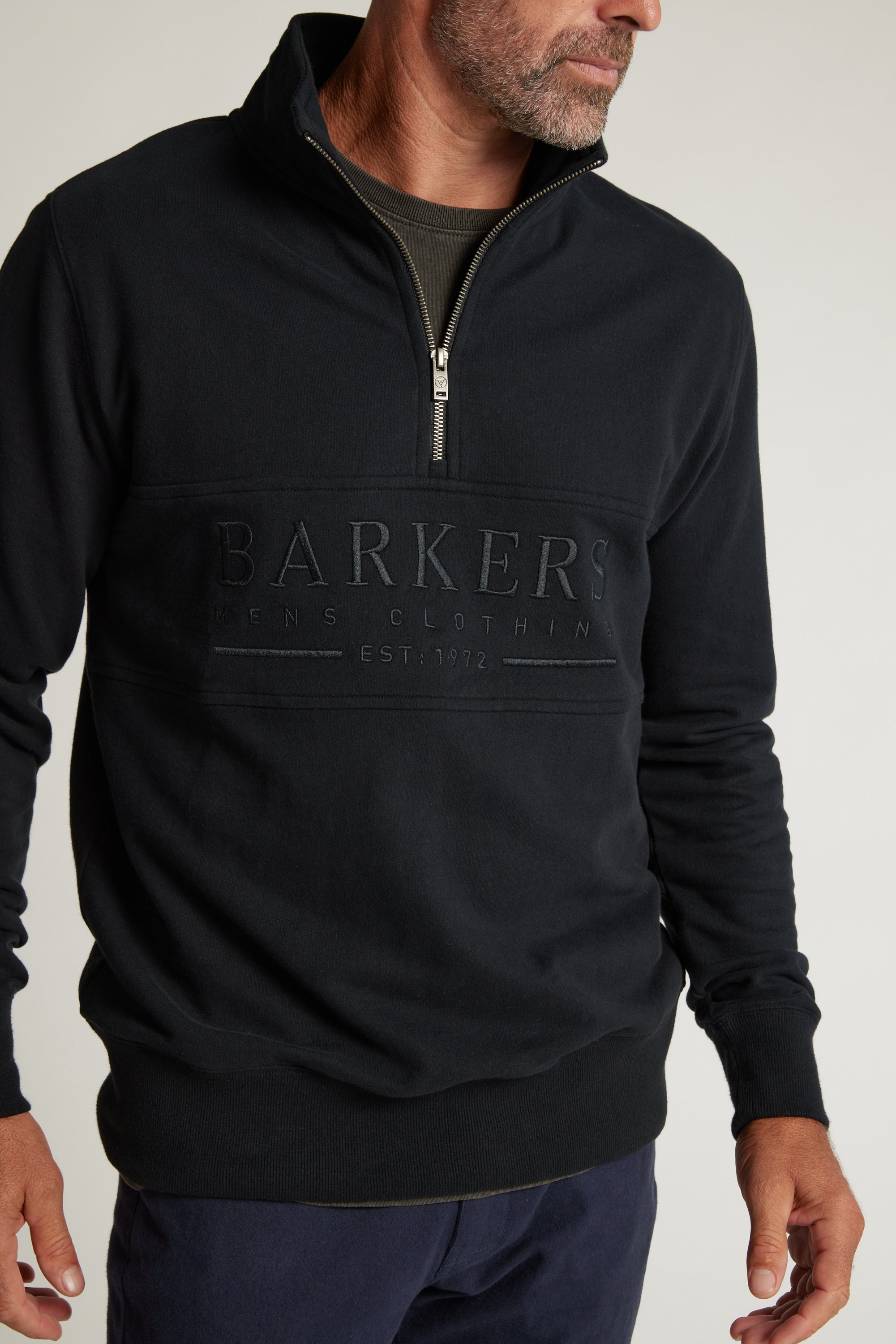 A man wears the Barkers Heritage 1/4 Zip sweater in navy, featuring “BARKERS MENS CLOTHING EST. 1972” embroidered on the front. He stands sideways with one hand in his pocket.