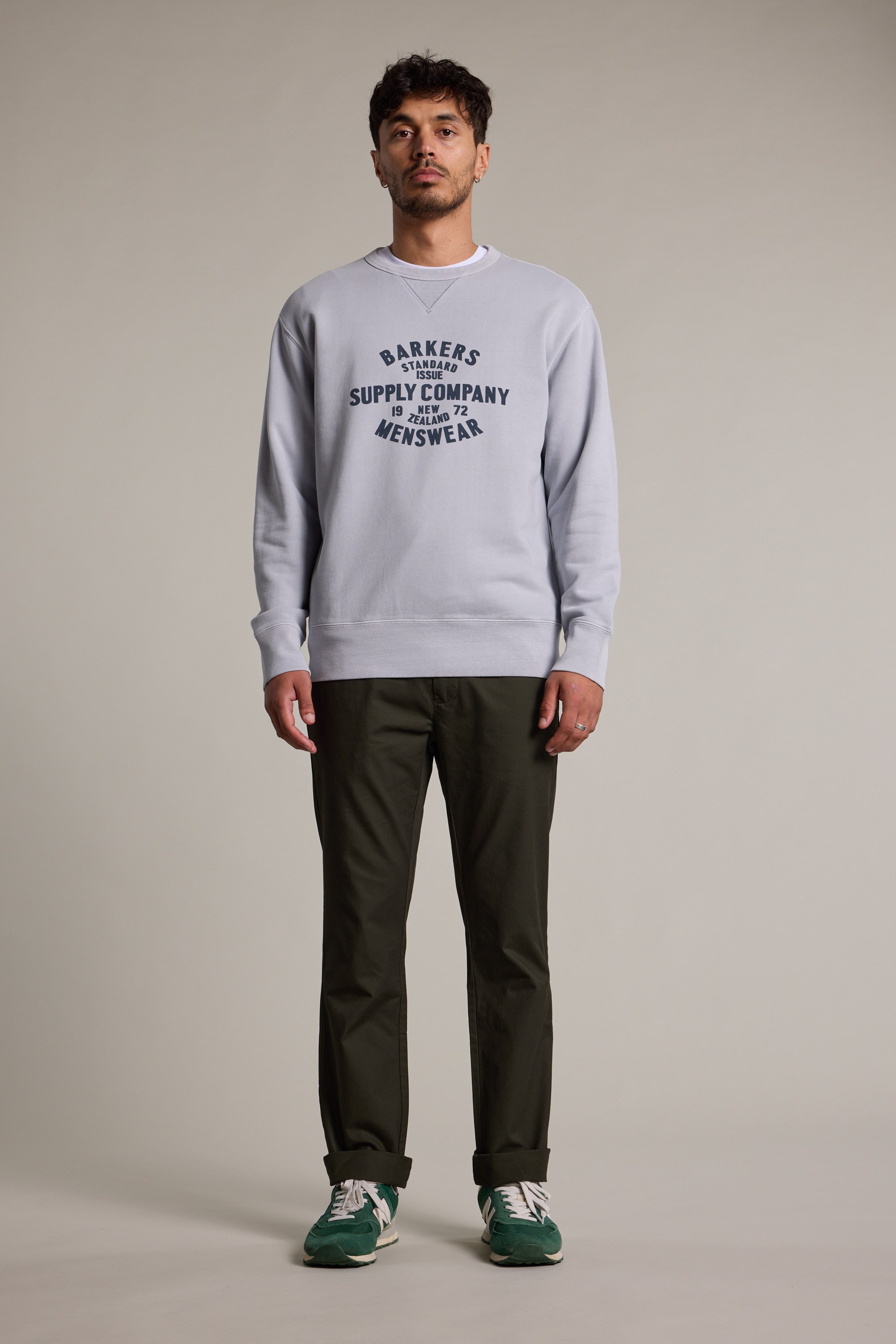A man faces forward in a Barkers Standard Issue Print Crew sweater in light gray, made from responsibly sourced cotton, styled with dark green pants and green sneakers. He stands against a plain neutral background.