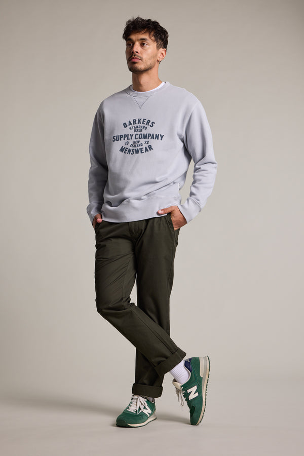 A man with short dark hair stands against a plain background wearing a Barkers Standard Issue Print Crew sweater in light gray fleece, dark green pants, and green New Balance sneakers. One hand is in his pocket as he looks slightly to the side.