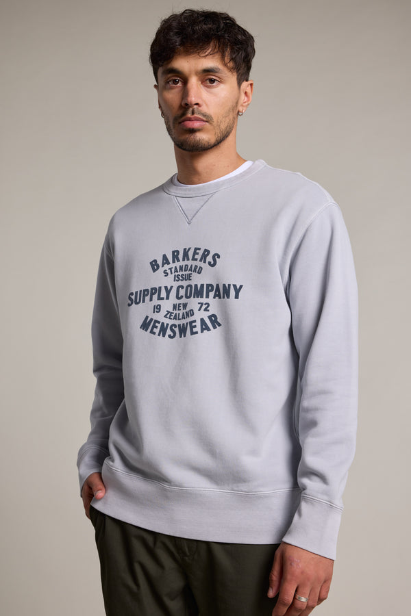 A man with short dark hair and facial hair wears the Barkers Standard Issue Print Crew, crafted from responsibly sourced cotton with bold front text. He stands against a plain light background, one hand in his pocket.