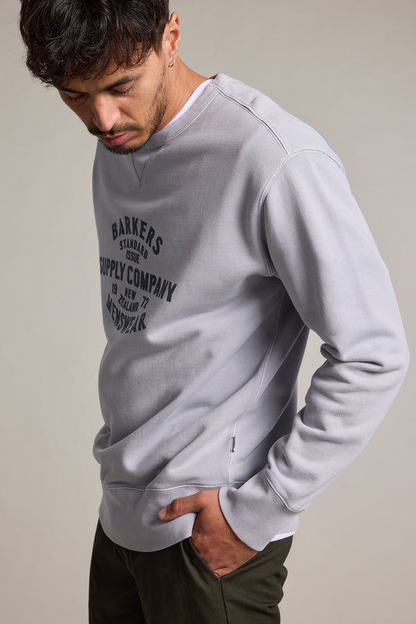 A man with short dark hair wears a Barkers Standard Issue Print Crew made from responsibly sourced cotton and dark green pants, looking down with one hand in his pocket against a plain background.