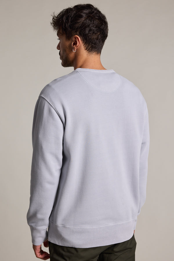 A man with short dark hair stands facing away from the camera, wearing a light gray Barkers Standard Issue Print Crew sweater made from responsibly sourced cotton and dark pants, set against a plain, light background.