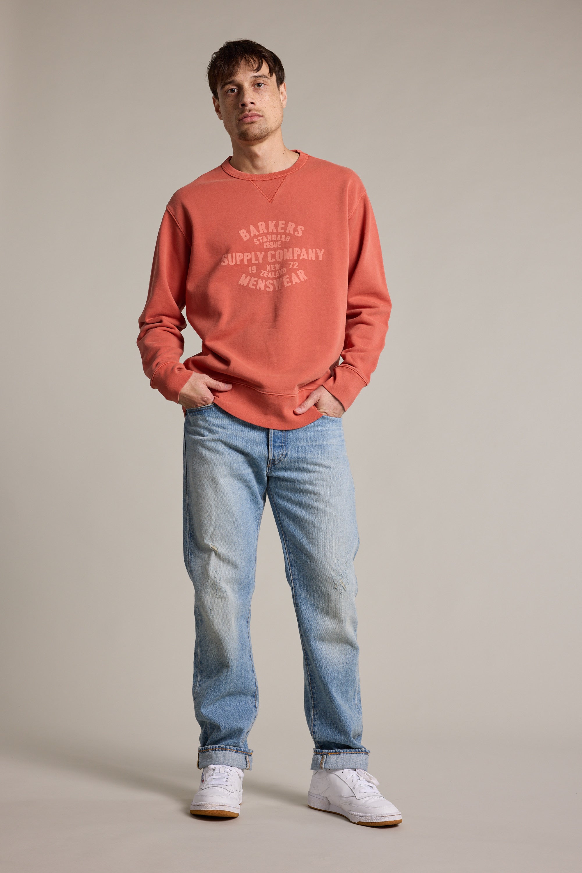 A man faces forward in a faded red Barkers Standard Issue Print Crew, made from responsibly sourced cotton, paired with light blue jeans and white sneakers. One hand is in his pocket; he poses neutrally against a plain beige background.