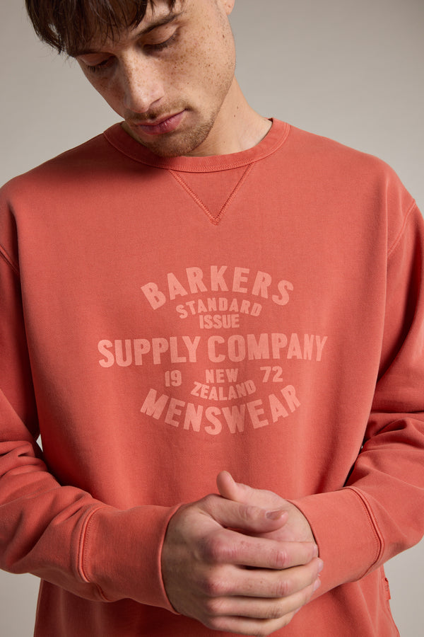 A person wearing a faded red Barkers Standard Issue Print Crew—made from responsibly sourced cotton and printed with "Barkers Supply Company Menswear, New Zealand, 1972"—looks down with hands clasped.