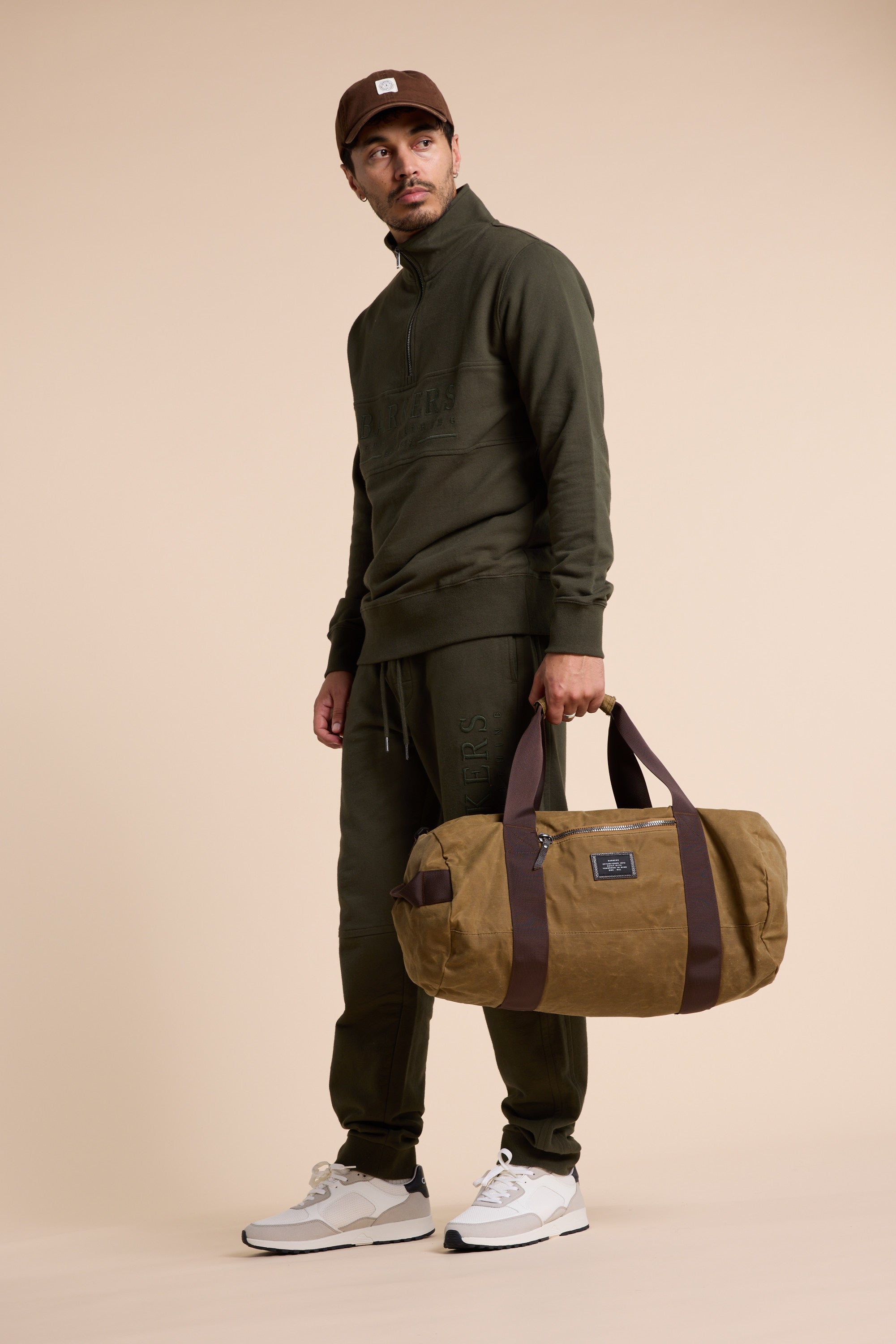 A man wearing a Barkers Heritage 1/4 Zip in olive green and brown cap stands holding a brown duffel bag. He pairs his outfit with white sneakers and looks slightly to the side against a plain beige background.