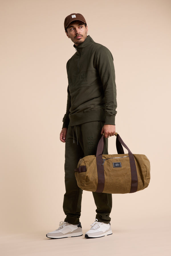 A man wearing a Barkers Heritage 1/4 Zip in olive green and brown cap stands holding a brown duffel bag. He pairs his outfit with white sneakers and looks slightly to the side against a plain beige background.