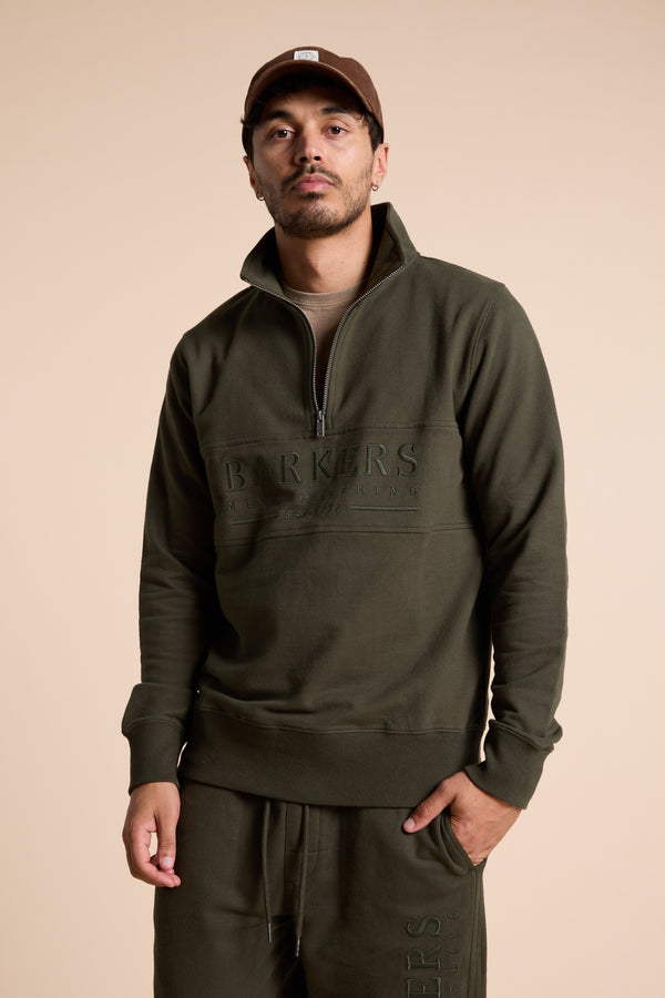 A man wears the Barkers Heritage 1/4 Zip in olive green with Barkers embossed on the front, matching joggers, and a brown cap, standing against a beige background with one hand in his pocket.