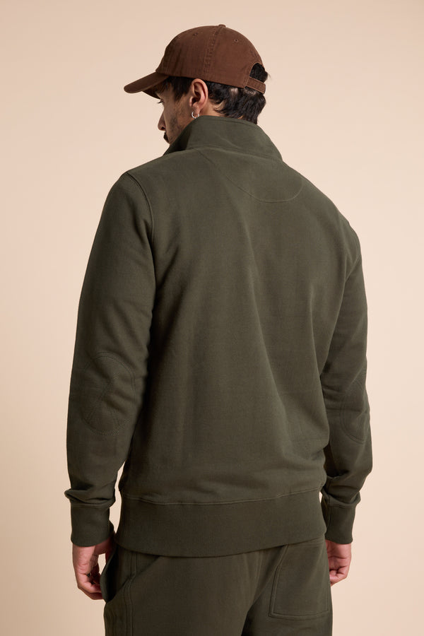 A man in an olive green Barkers Heritage 1/4 Zip by Barkers and matching pants faces away from the camera, wearing a brown baseball cap against a plain beige background.
