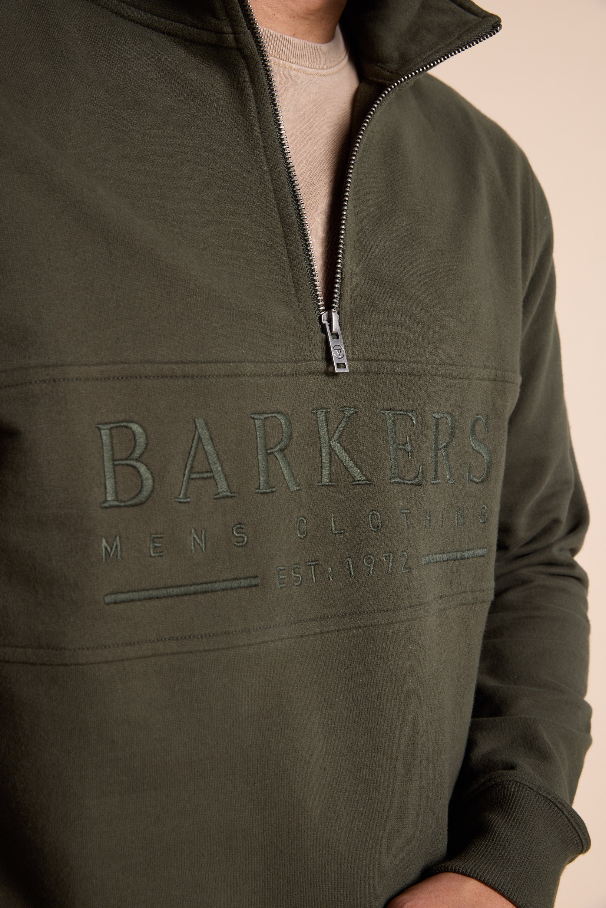 A person wears a dark green Barkers Heritage 1/4 Zip sweater by Barkers over a beige t-shirt. "Barkers Mens Clothing Est. 1972" is embroidered on the front, and the person's face is not visible.