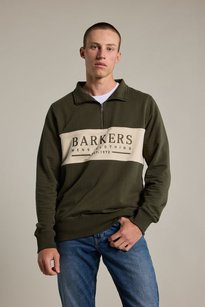 A young man in a khaki green Barkers Heritage 1/4 Zip sweater and blue jeans stands against a plain light background, looking at the camera.