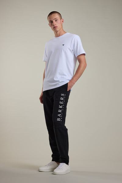 A man stands in a relaxed pose wearing a plain white T-shirt, Barkers Organic Classic Print Track Pant with "BARKERS" printed down the leg in white, and white sneakers against a neutral background.