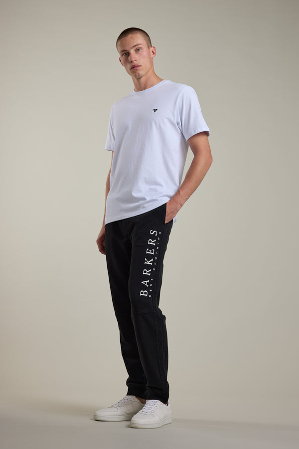 A man stands in a relaxed pose wearing a plain white T-shirt, Barkers Organic Classic Print Track Pant with "BARKERS" printed down the leg in white, and white sneakers against a neutral background.