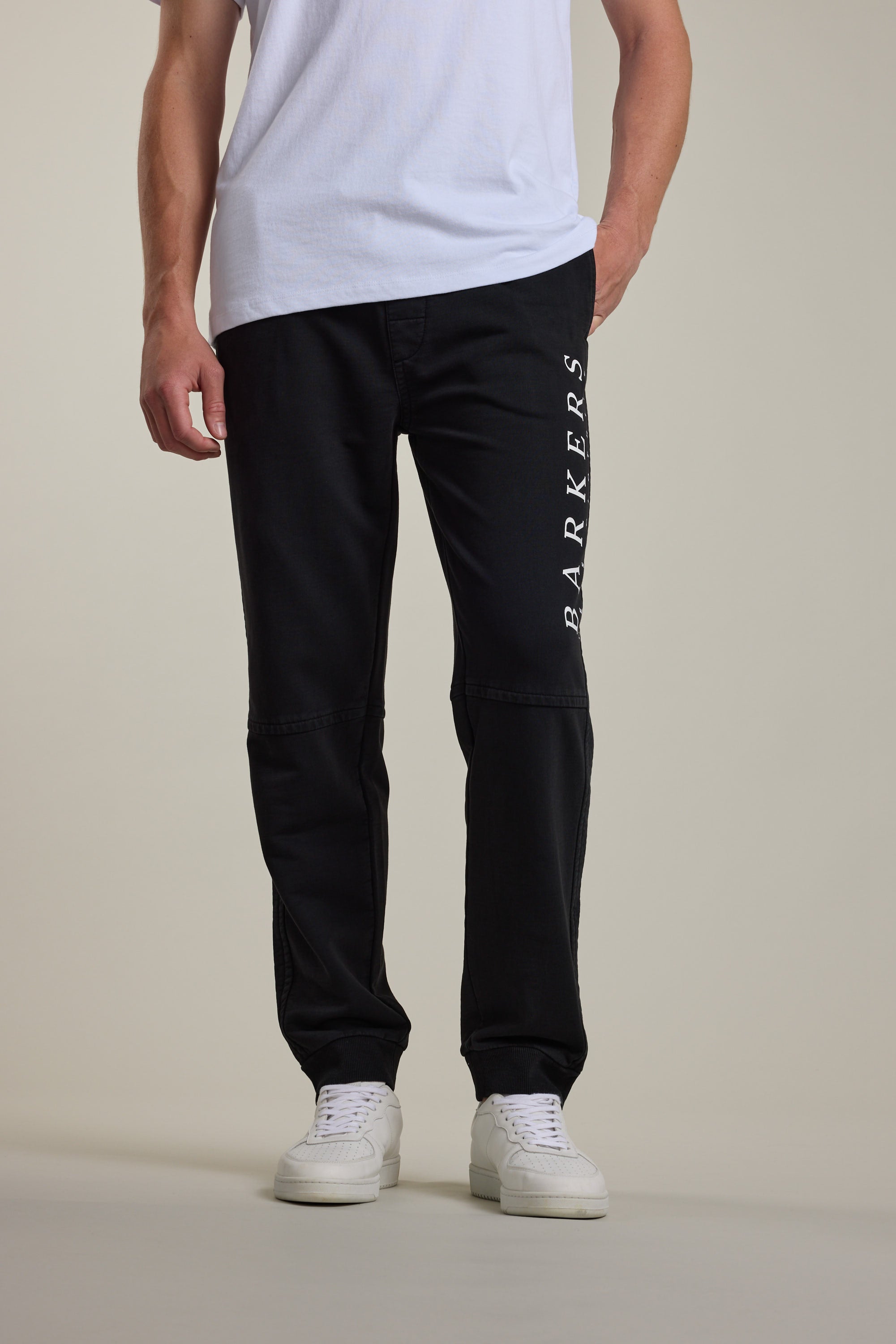 A person wearing a white t-shirt, Barkers Organic Classic Print Track Pant with "BARKERS" printed vertically on one leg, and white sneakers stands against a plain background.