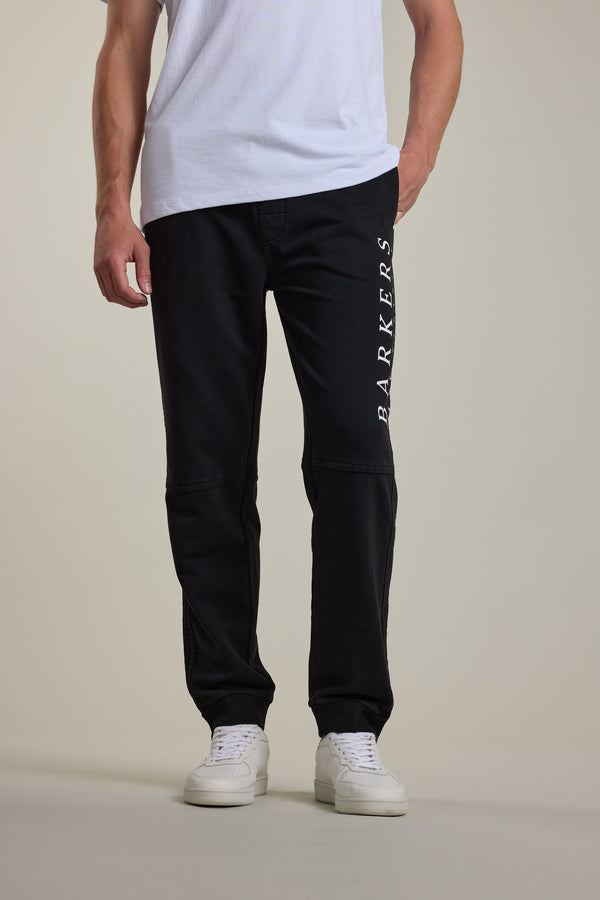 A person wearing a white t-shirt, Barkers Organic Classic Print Track Pant with "BARKERS" printed vertically on one leg, and white sneakers stands against a plain background.