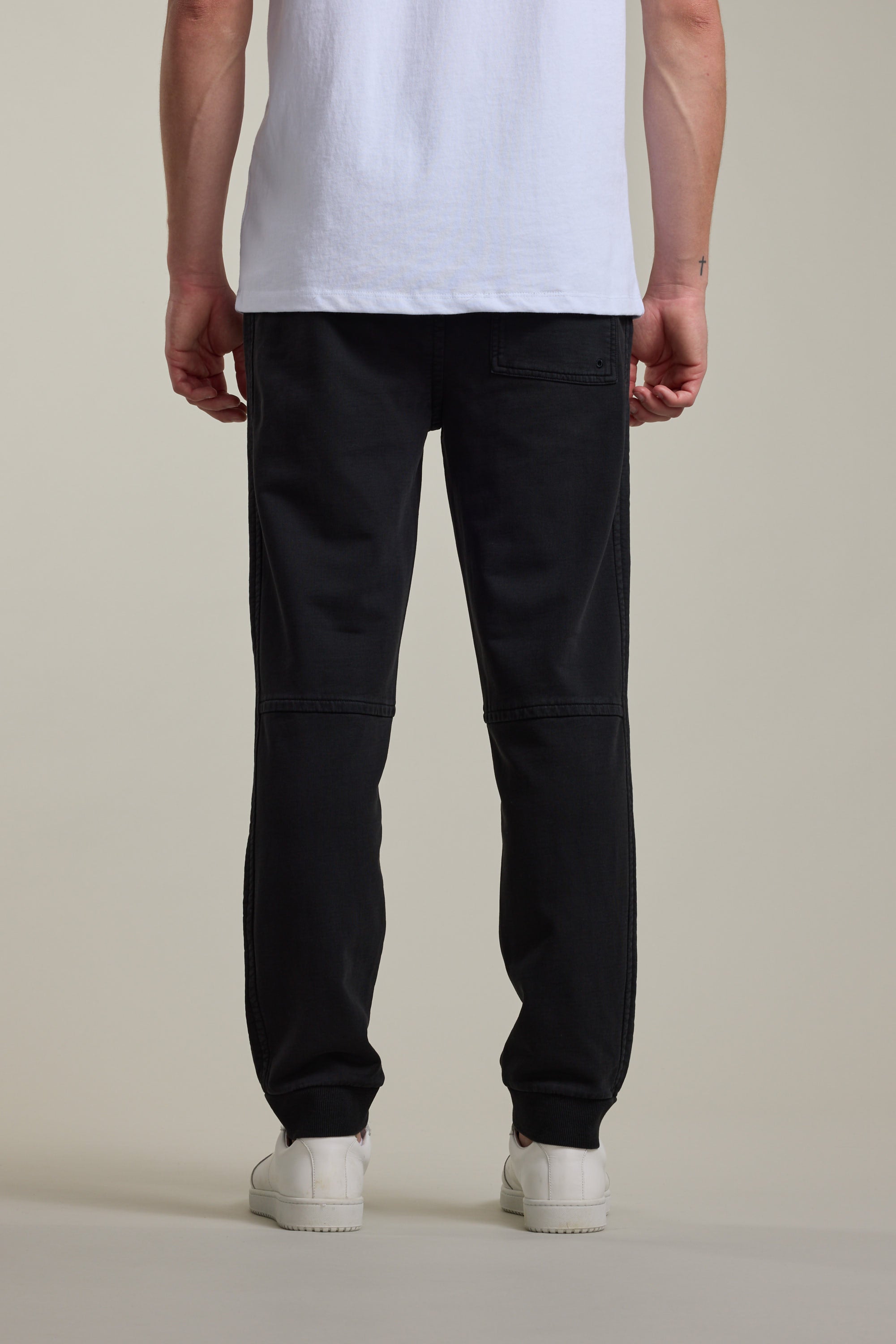 A person stands facing away, wearing Barkers Organic Classic Print Track Pant, a white t-shirt, and white sneakers against a plain light background, shown from shoulders down.