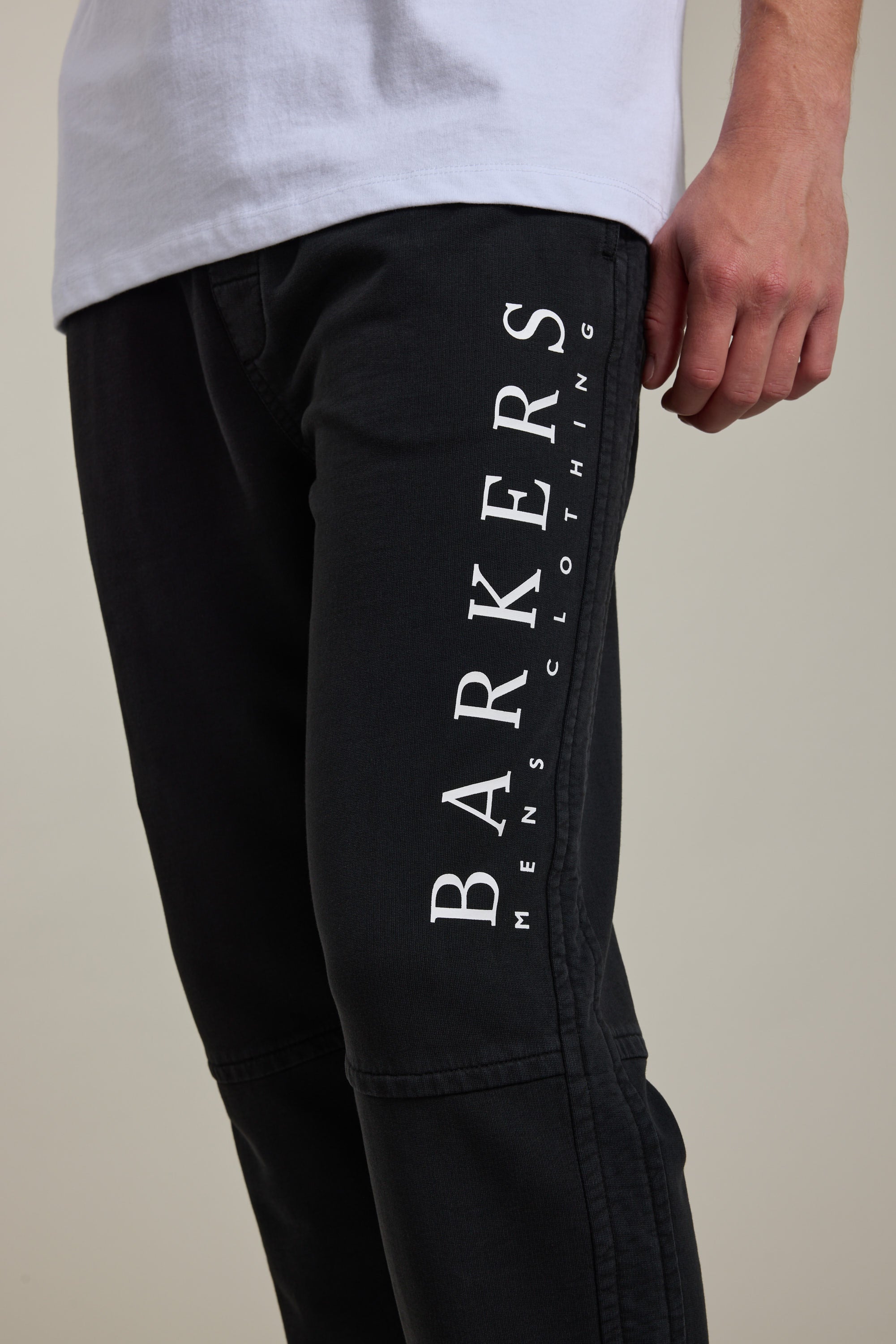 A person wearing Barkers Organic Classic Print Track Pants in black, featuring “BARKERS MENS CLOTHING” printed vertically in white on the leg, paired with a white shirt. Only the lower torso and hand are visible.