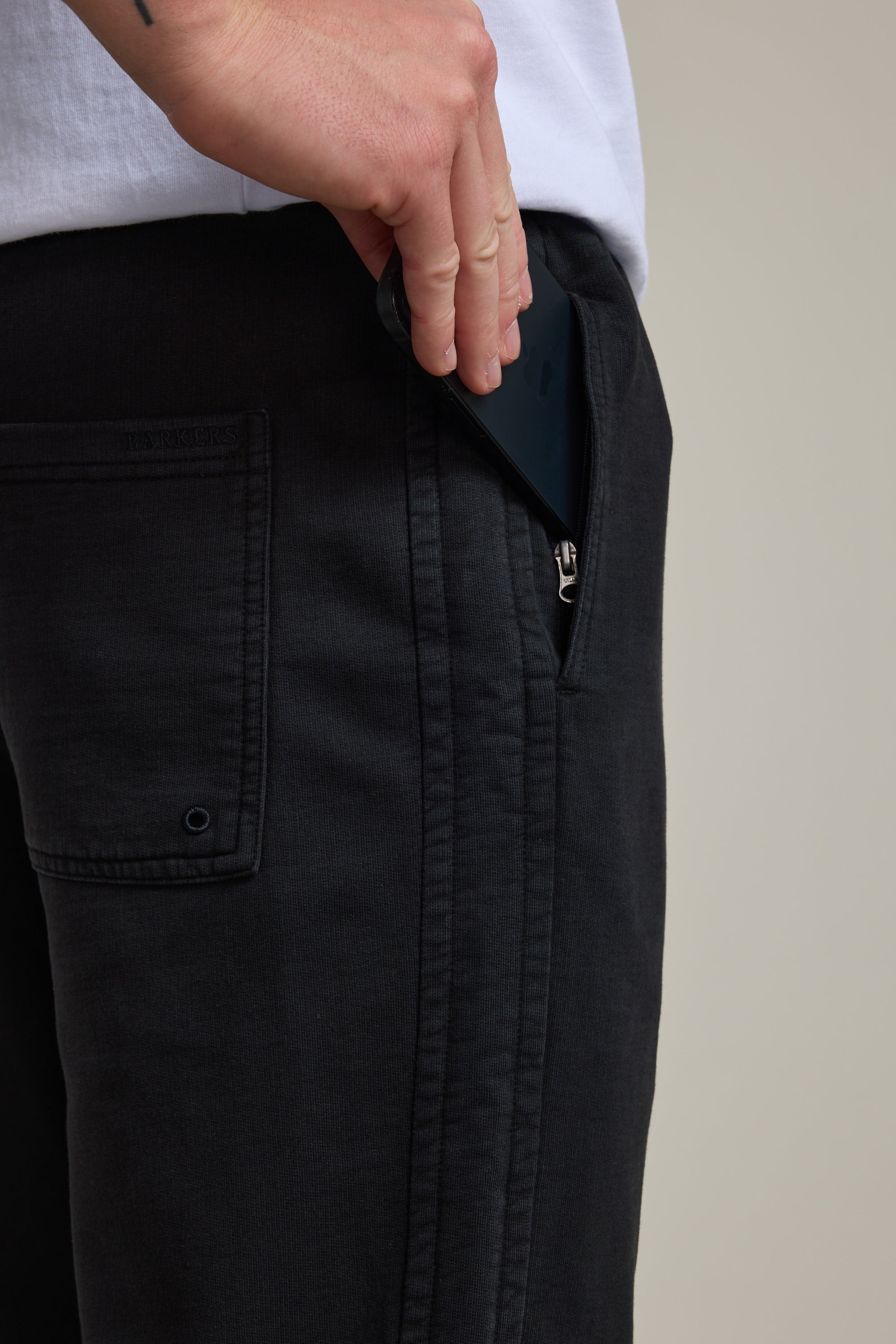 A person wearing Barkers Organic Classic Print Track Pant is placing a black smartphone into the zippered side pocket, with only their lower torso and hand visible.