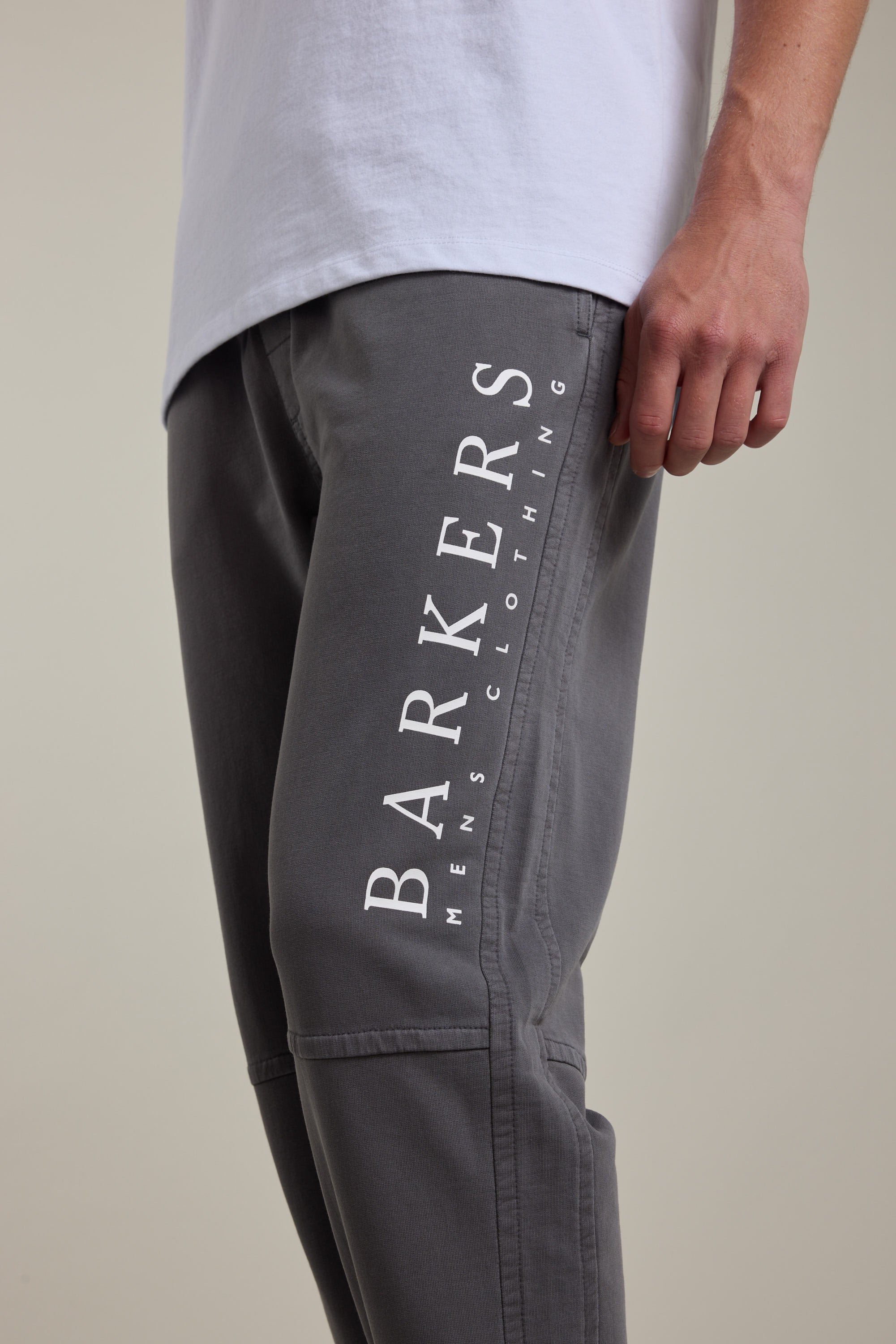 A person is shown from the waist down wearing Barkers Organic Classic Print Track Pant in grey, featuring “BARKERS MENS CLOTHING” printed vertically in white on the thigh, paired with a white shirt.