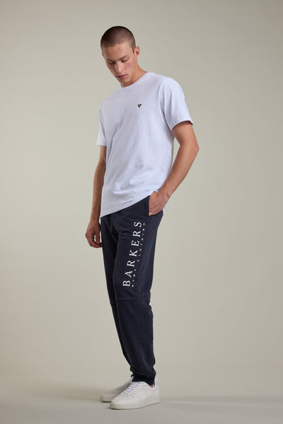 A man stands against a plain background wearing a white t-shirt, Barkers Organic Classic Print Track Pant with "BARKERS" down the leg, and white sneakers. One hand is in his pocket as he looks down.