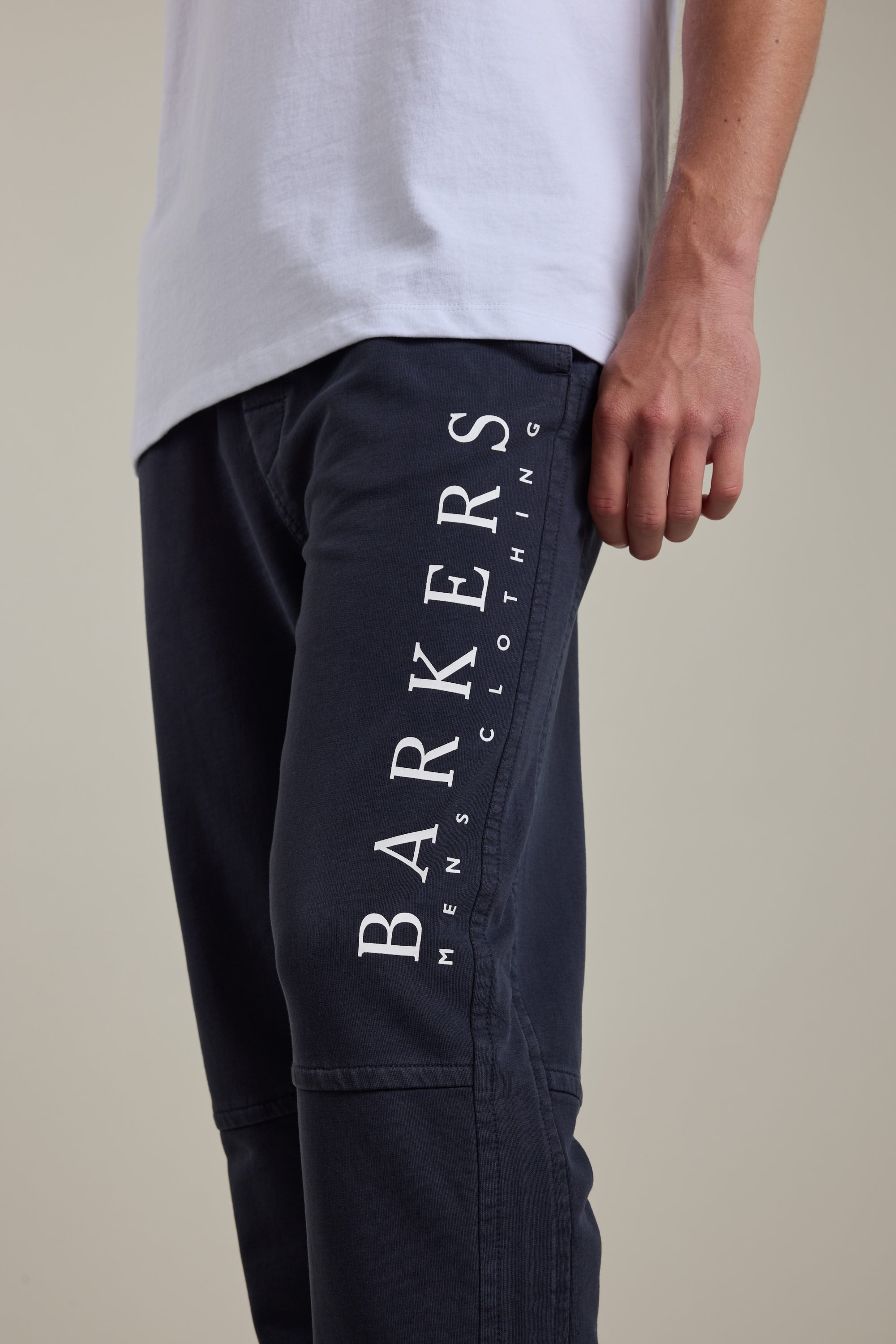 Someone wearing Barkers' Organic Classic Print Track Pant in dark blue, with “BARKERS MENS CLOTHING” printed vertically in white on the thigh, paired with a white tee; only the lower torso and arm are shown.