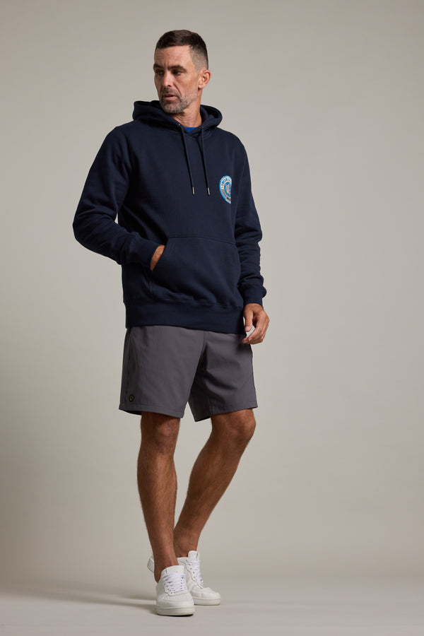 A man stands against a plain background wearing a navy Swordfish Mojo Logo Hoody, gray shorts, and white sneakers. His left hand is in his pocket and he looks to the side with a neutral expression.