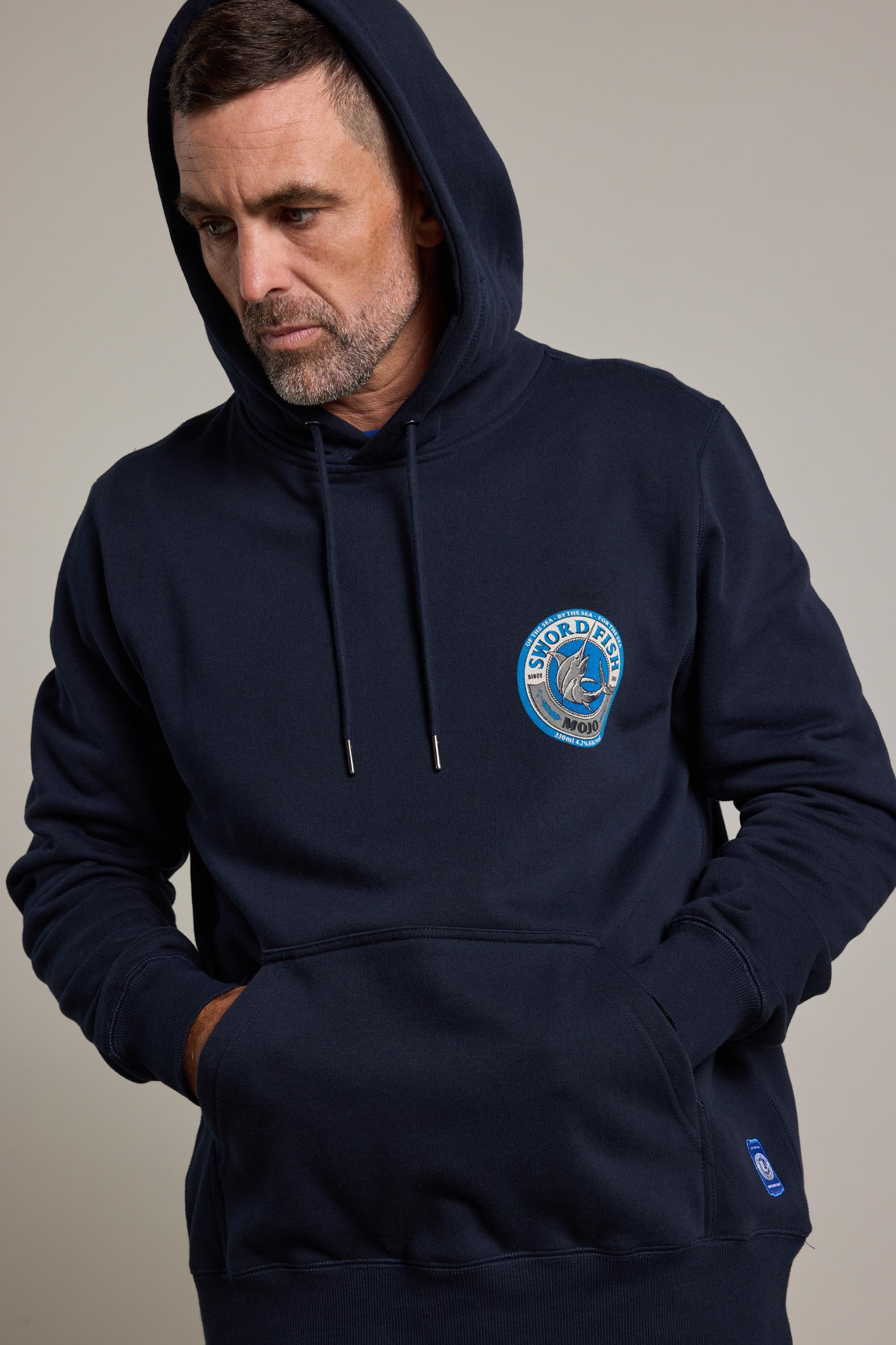 A man wears the navy blue Swordfish Mojo Logo Hoody, made from responsibly sourced cotton, featuring a circular chest logo and a small pocket tag, with hands in the front pocket, looking down and to the side.