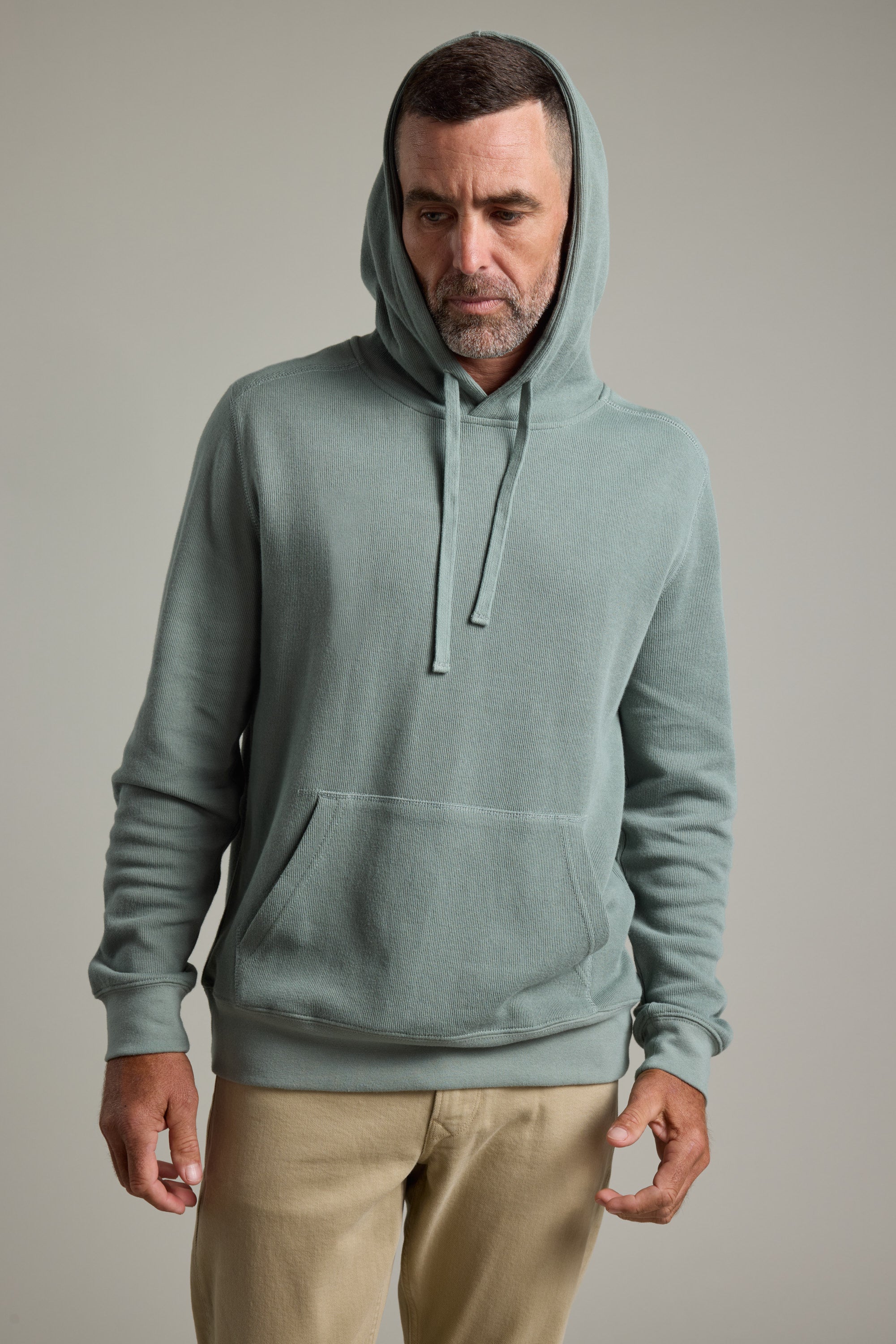 A man with short hair and a trimmed beard wears the Barkers Inlet Hoody in light green and beige pants. He stands against a plain background, looking down with a neutral expression and his hands relaxed at his sides.