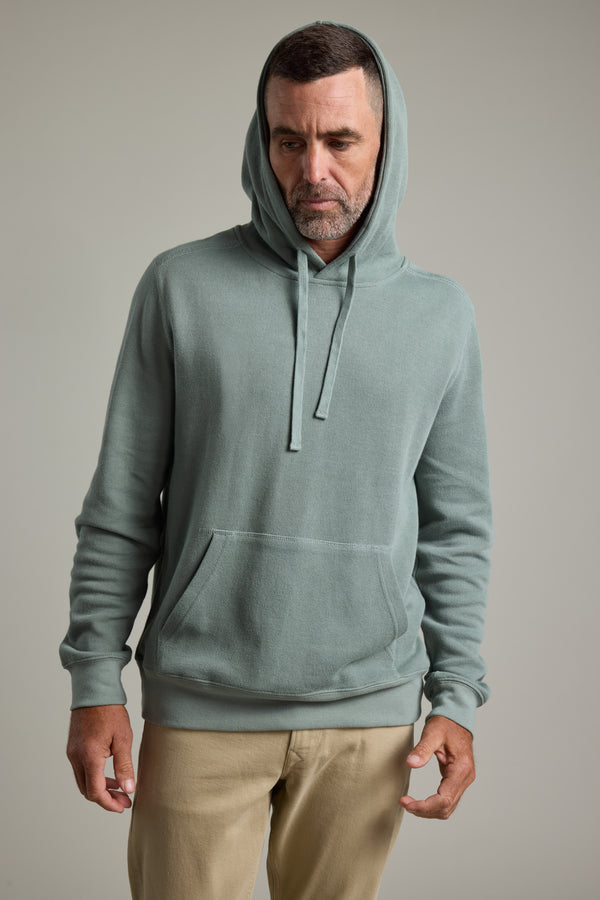 A man with short hair and a trimmed beard wears the Barkers Inlet Hoody in light green and beige pants. He stands against a plain background, looking down with a neutral expression and his hands relaxed at his sides.