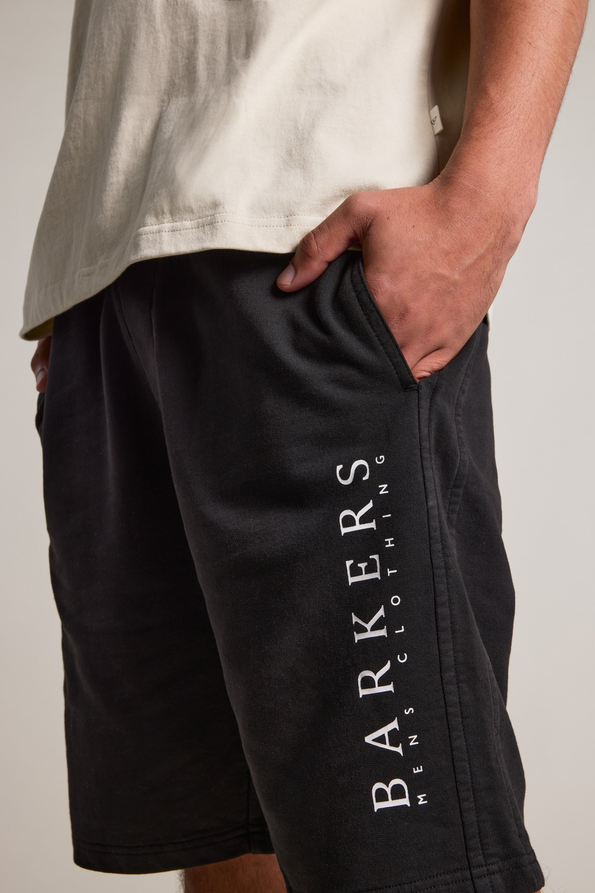 A person wearing a light-colored T-shirt and relaxed fit Barkers Organic Classic Print Track Shorts, with one hand in their pocket. The black shorts display "BARKERS" printed vertically on the side.