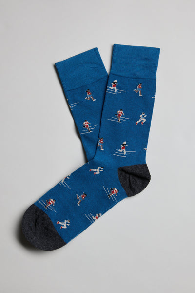The Barkers Kudos Runner Sock is a pair of blue cotton socks with dark gray toes, featuring small skier motifs in various poses, displayed flat on a light gray background.