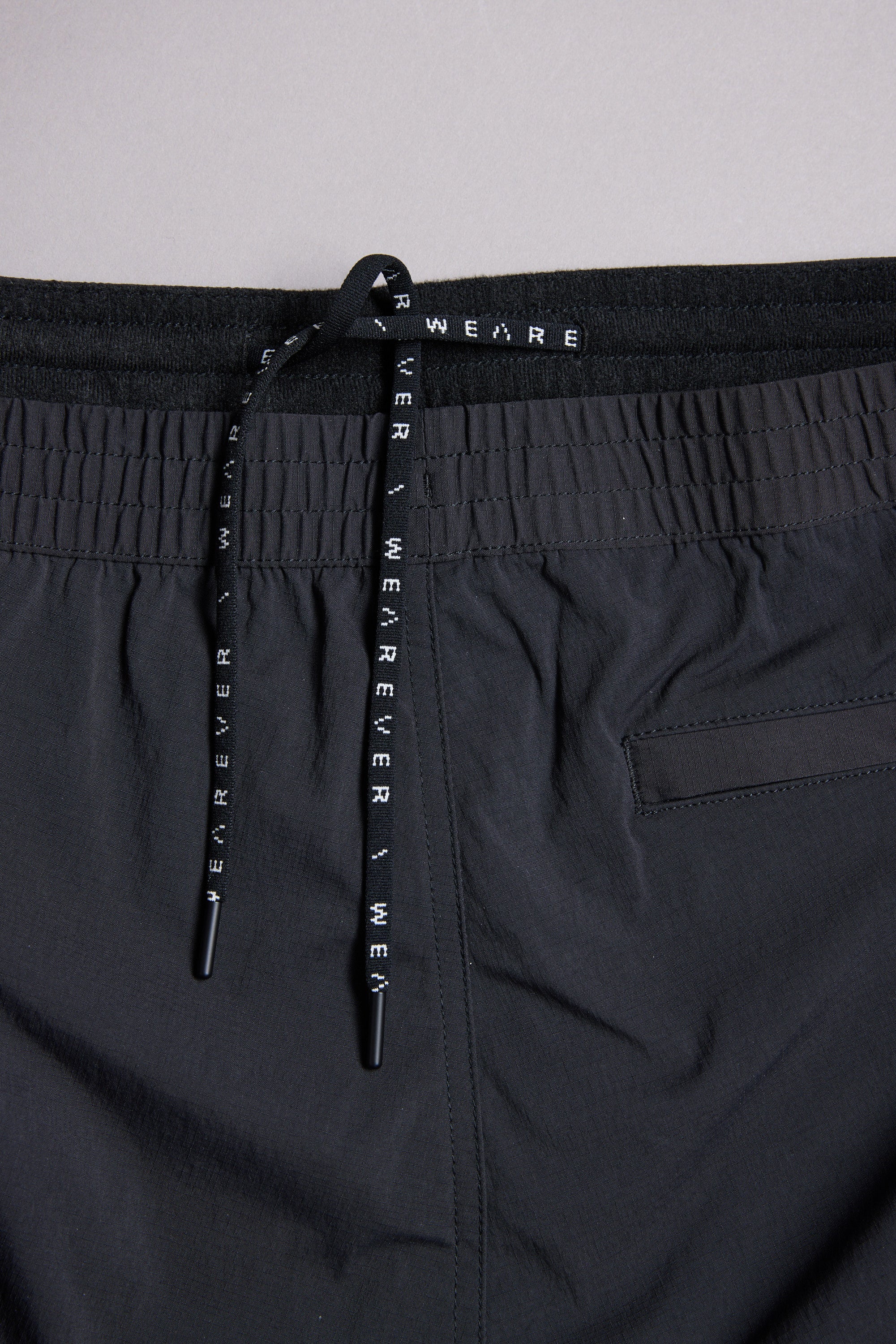 Close-up of the WearEver Transit Pant's black waistband with a drawstring featuring repeating WEAR EVER text. A subtle horizontal back pocket seam is partially visible on the right side.