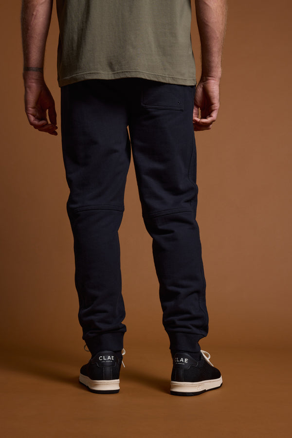 A person faces away from the camera against a brown background, wearing a green t-shirt, black sneakers with white soles, and Barkers Heritage Track Pant by Barkers for a classic yet relaxed style.