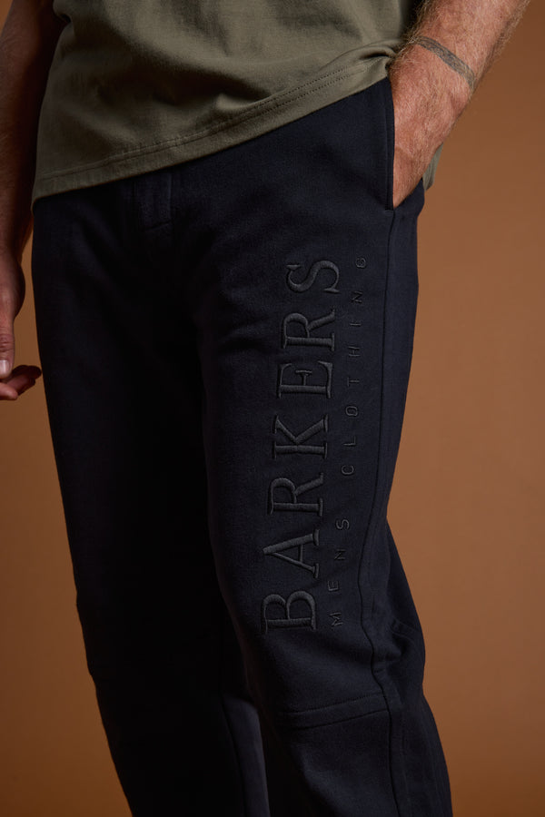 A person in Barkers Heritage Track Pant (black, with BARKERS MENS CLOTHING embroidery) and an olive green t-shirt stands against a brown background.