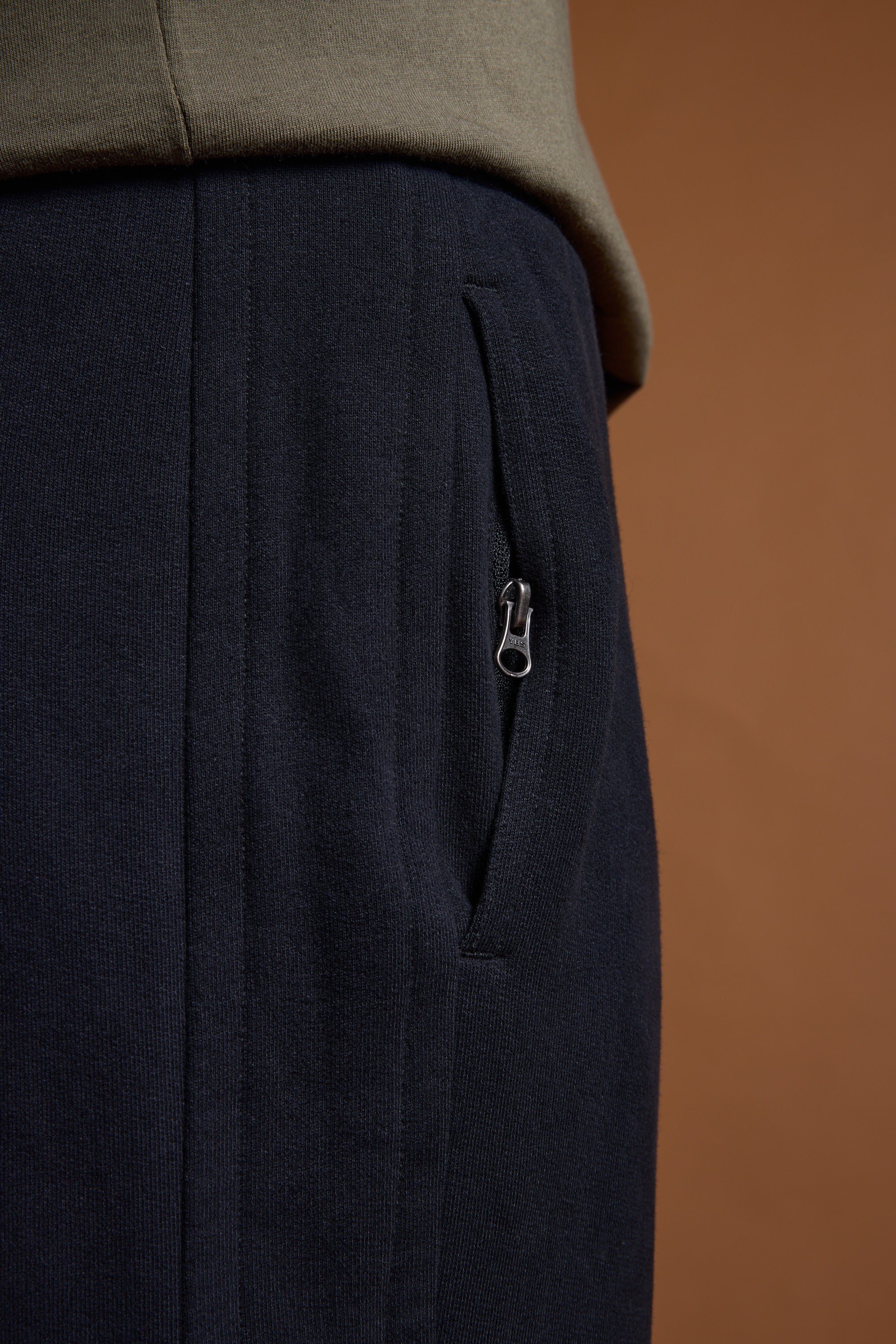 Close-up of a person wearing Barkers Heritage Track Pant by Barkers in dark navy with a zippered pocket, paired with an olive green top against a brown background.