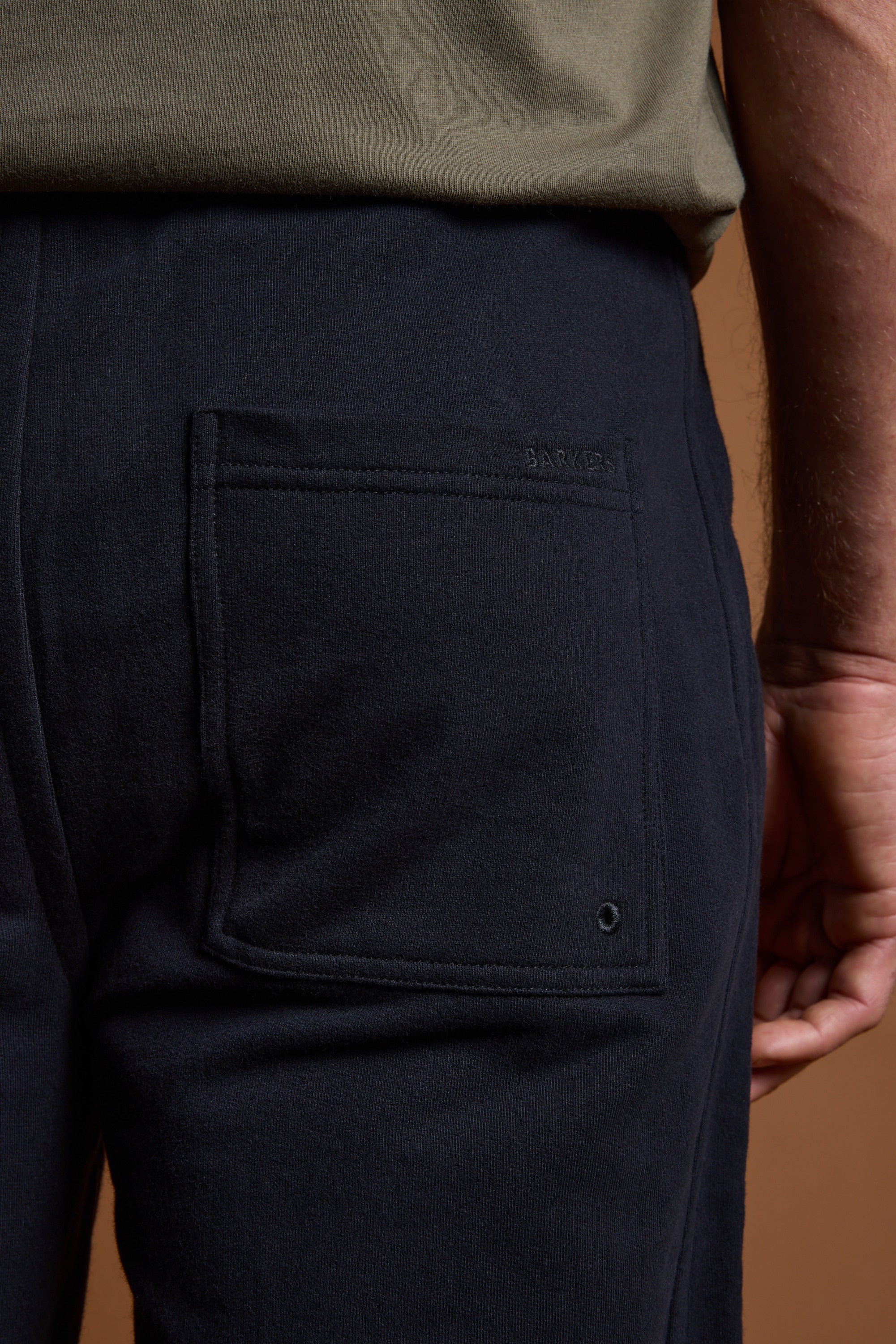 A person wears Barkers Heritage Track Pant in dark navy with a visible back pocket, paired with an olive green shirt. Their right arm rests by their side against a brown background. Brand: Barkers.