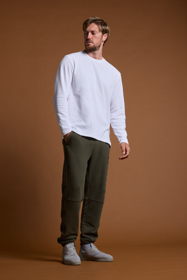 A man with light brown hair and a beard stands against a brown background, wearing a white long-sleeve shirt, Barkers Heritage Track Pant by Barkers in olive green, and light gray sneakers; his left hand is partly in his pocket.