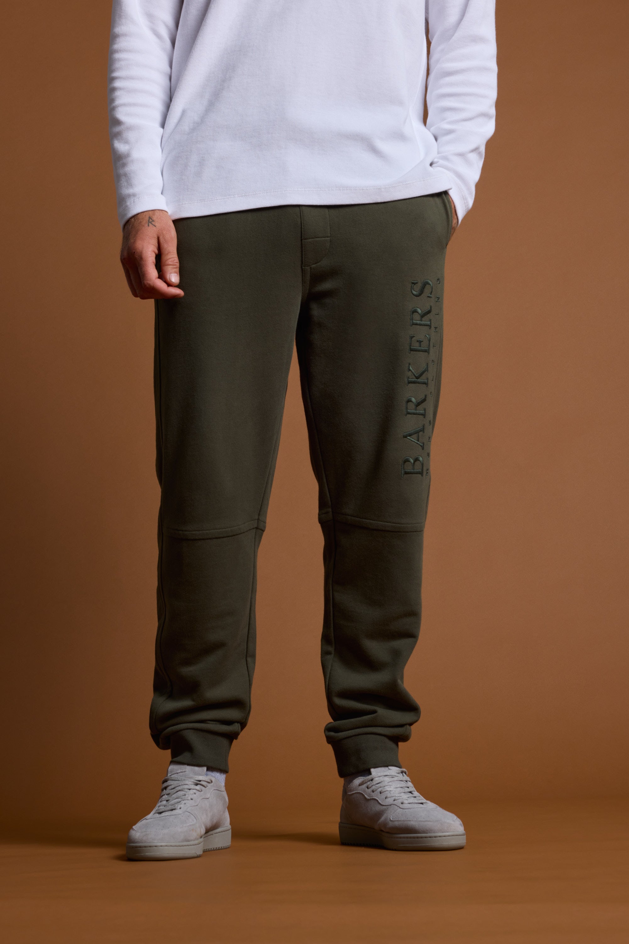 Someone stands against a brown background, dressed in a white long-sleeve shirt, Barkers Heritage Track Pant in dark green with "BARKERS" along the leg, and light gray sneakers.