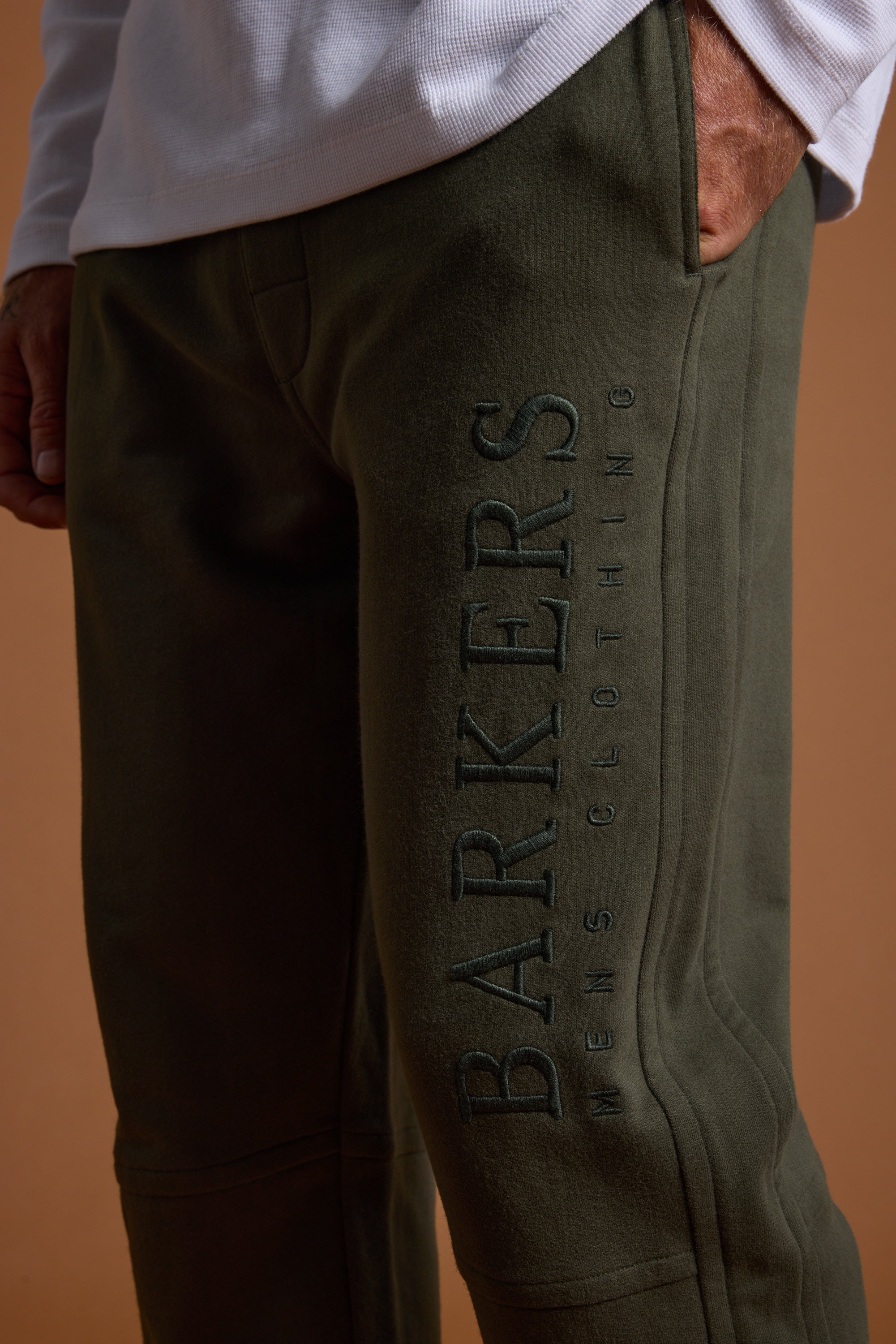 A close-up shows a person wearing olive green Barkers Heritage Track Pant by Barkers, with "BARKERS MENS CLOTHING" embroidered vertically on the left leg, paired with a white long-sleeve shirt and one hand in their pocket.