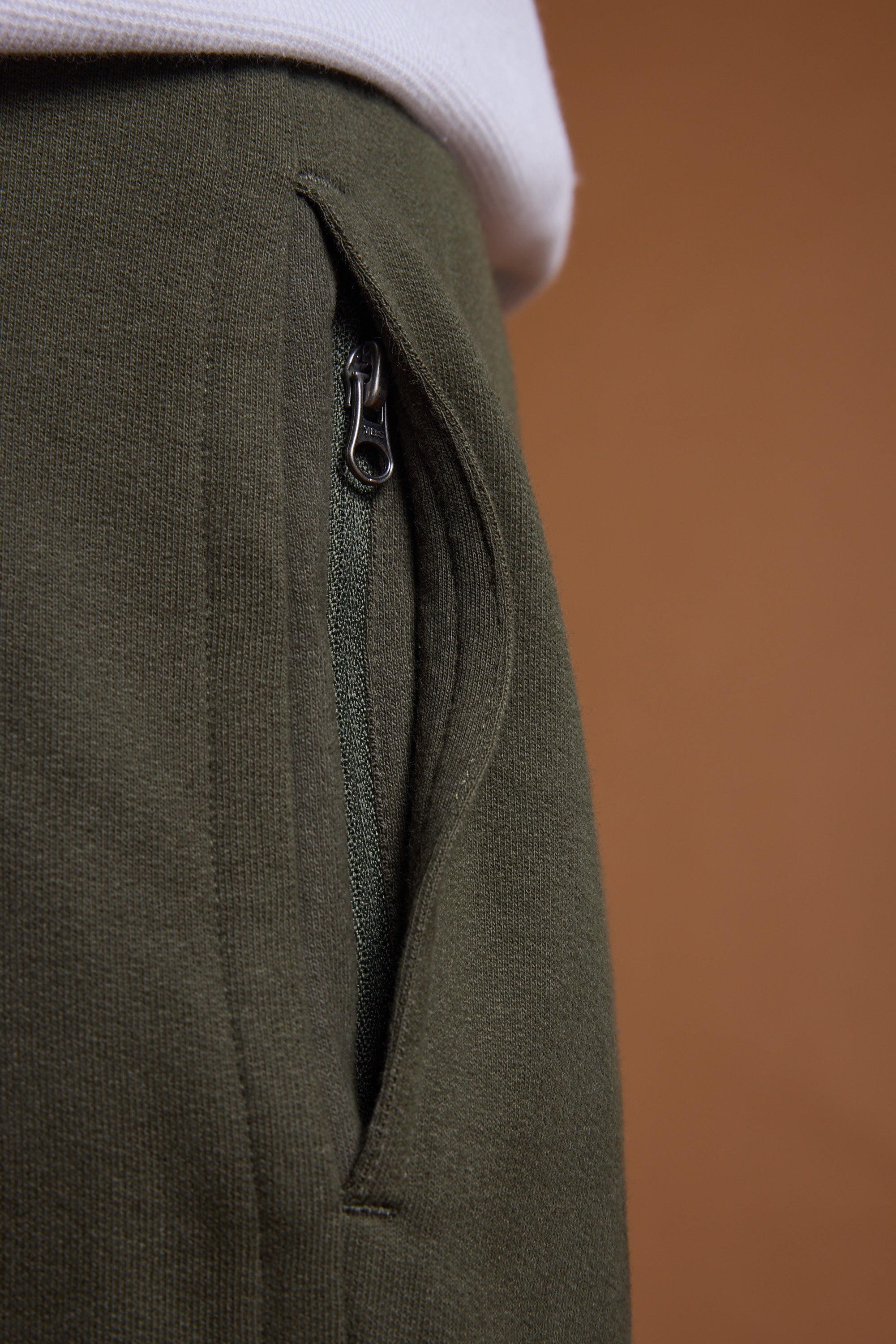 Close-up of a green zippered pocket on the Barkers Heritage Track Pant by Barkers, with part of a white shirt at the top set against a brown background.