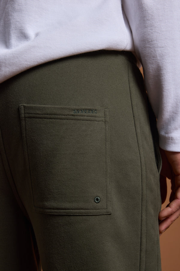 Close-up of a person wearing Barkers Heritage Track Pant in olive green, featuring a back pocket with small grommet and "BARKERS" embroidery above it, styled with a white textured shirt.