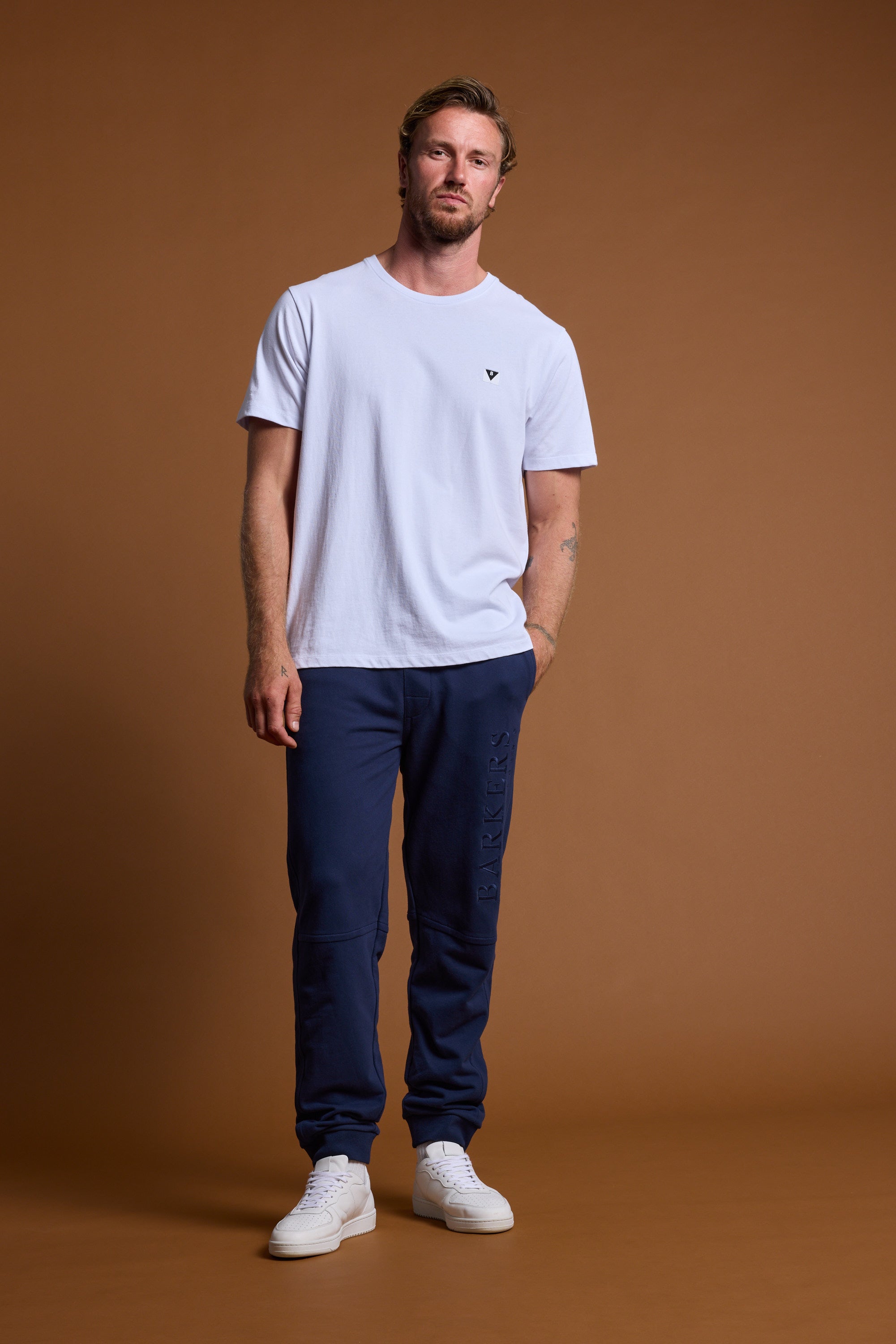 A man with short hair and a beard stands against a brown backdrop, wearing a white t-shirt, white sneakers, and navy Barkers Heritage Track Pants by Barkers, posing in a relaxed stance with one hand in his pocket.