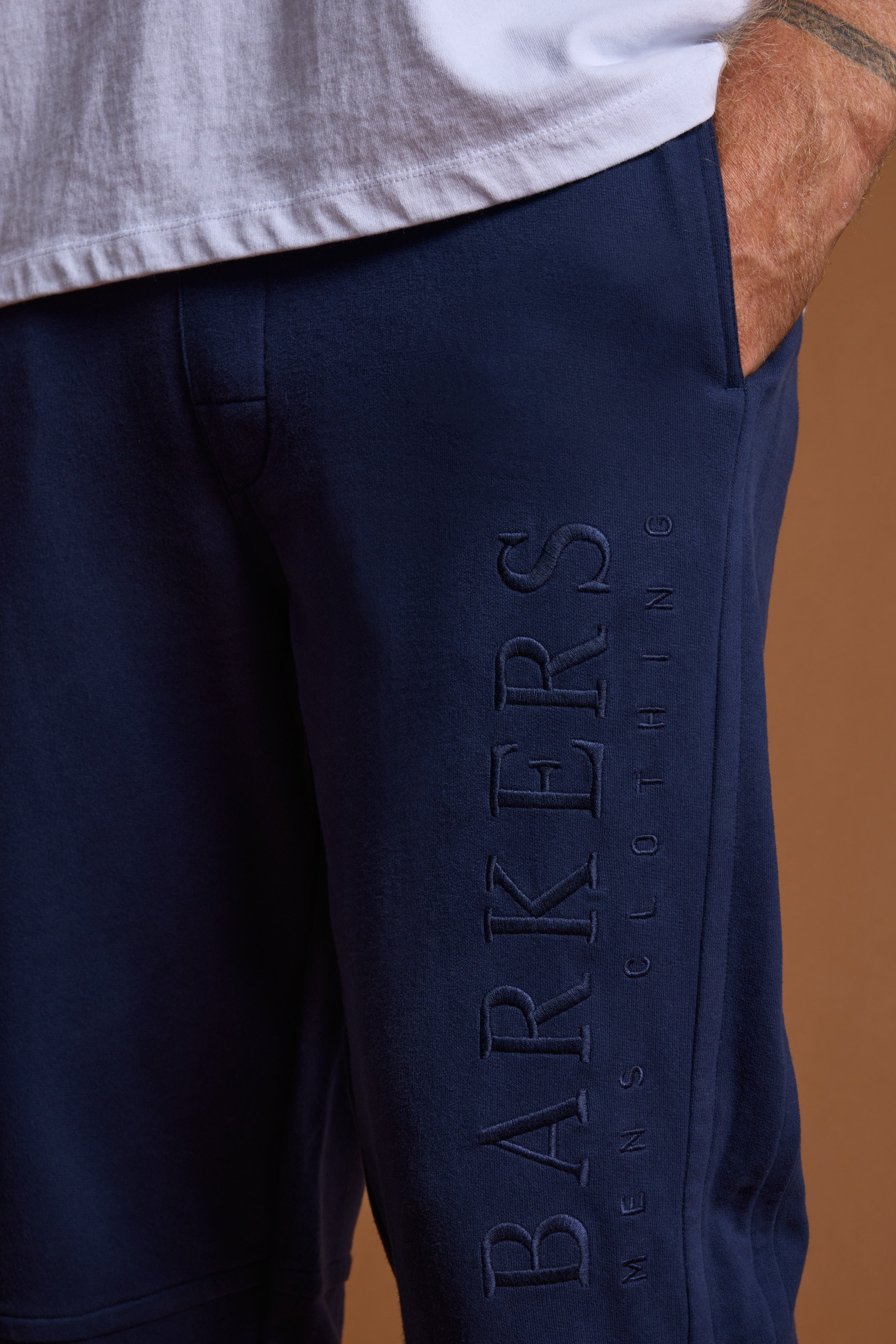 A person wears Barkers Heritage Track Pant in navy, featuring BARKERS MENS CLOTHING embossed vertically on the leg, paired with a partially tucked-in white shirt and one hand in the pocket.