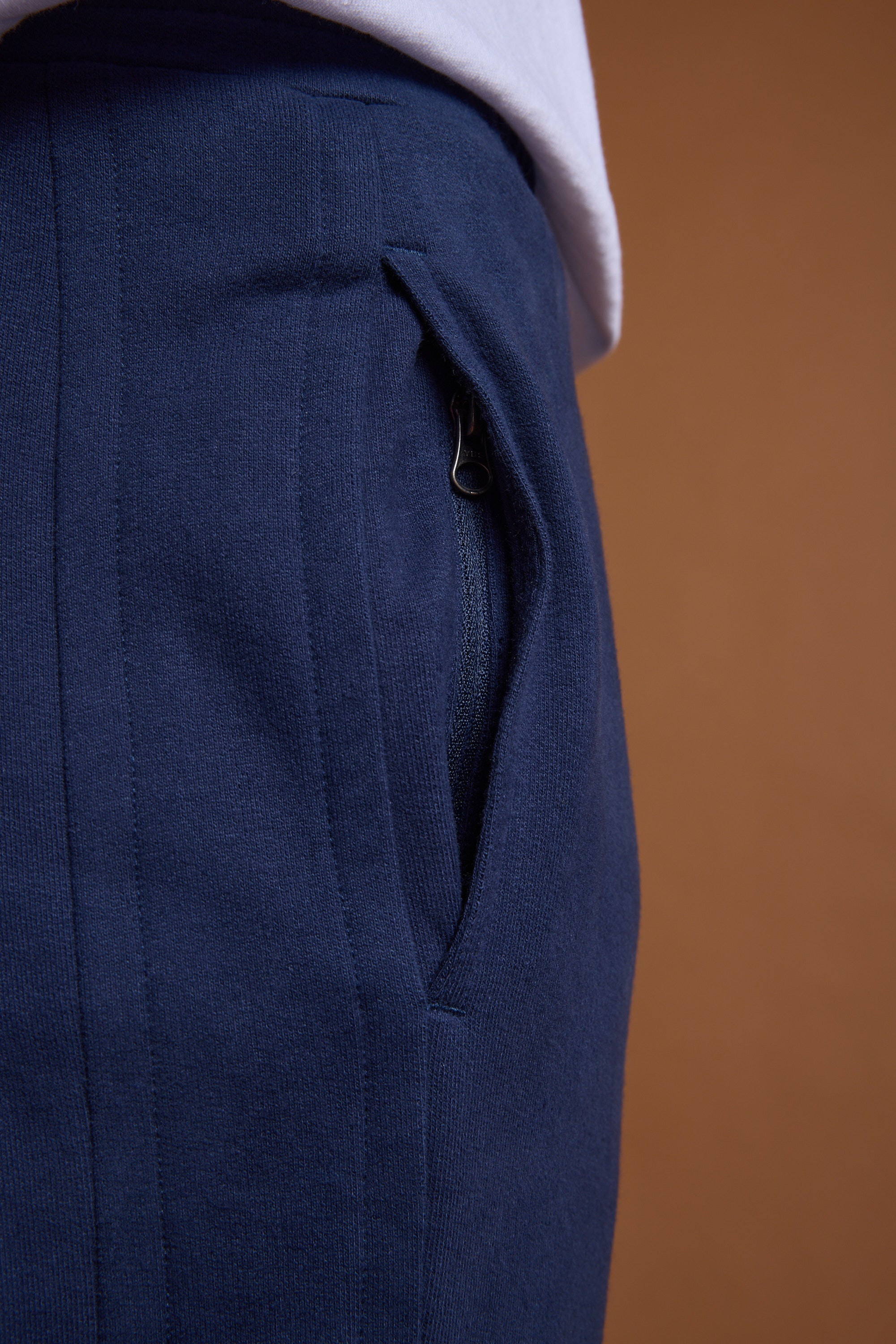 Close-up of a person wearing Barkers Heritage Track Pant in navy blue organic cotton with a zippered pocket, styled with a slightly tucked-in white shirt, by Barkers, against a brown background.