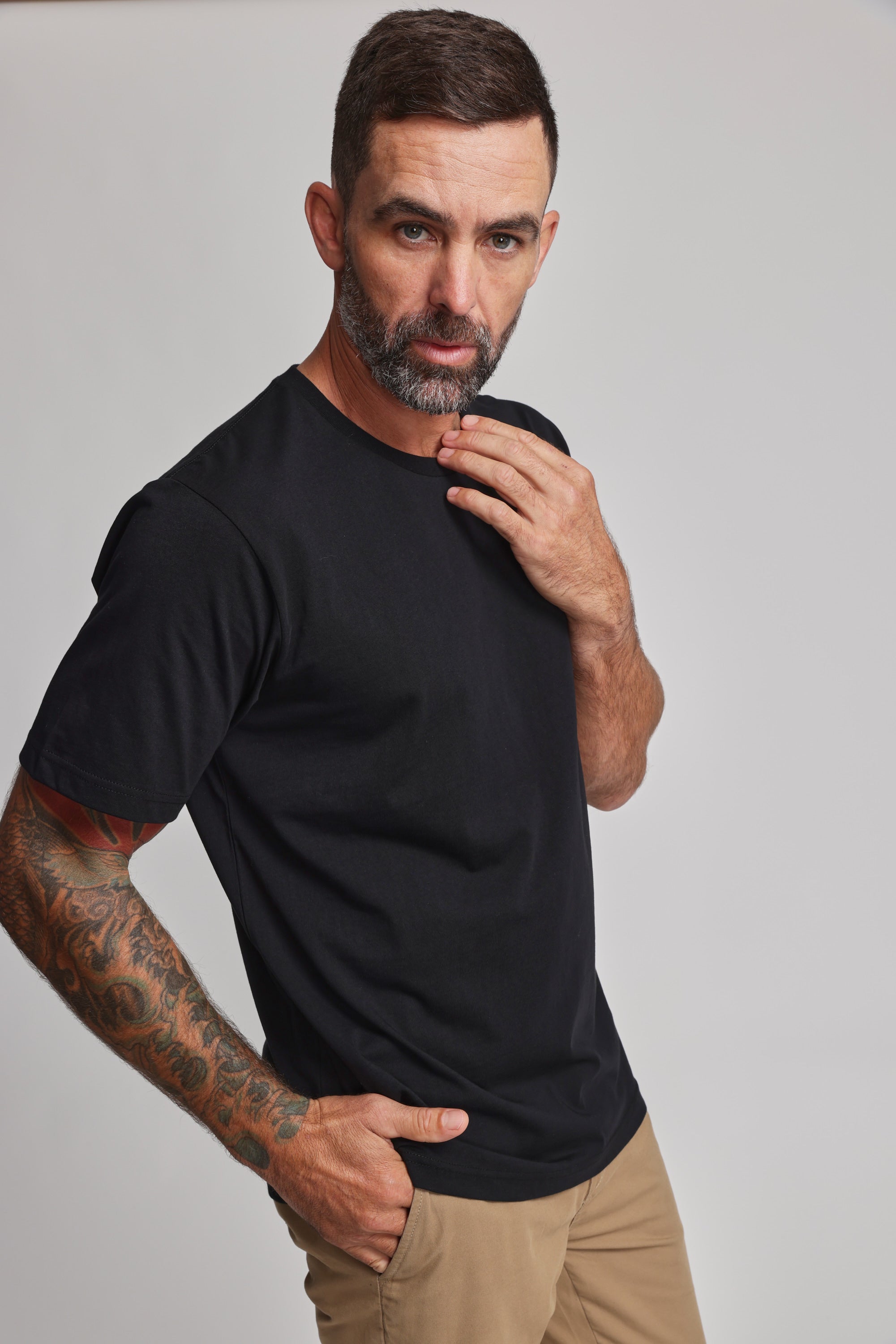 A man with short dark hair, a beard, and tattoos on his left arm poses against a plain background wearing the Barkers Organic Crew Tee in black with beige pants, one hand on his chin and the other in his pocket.