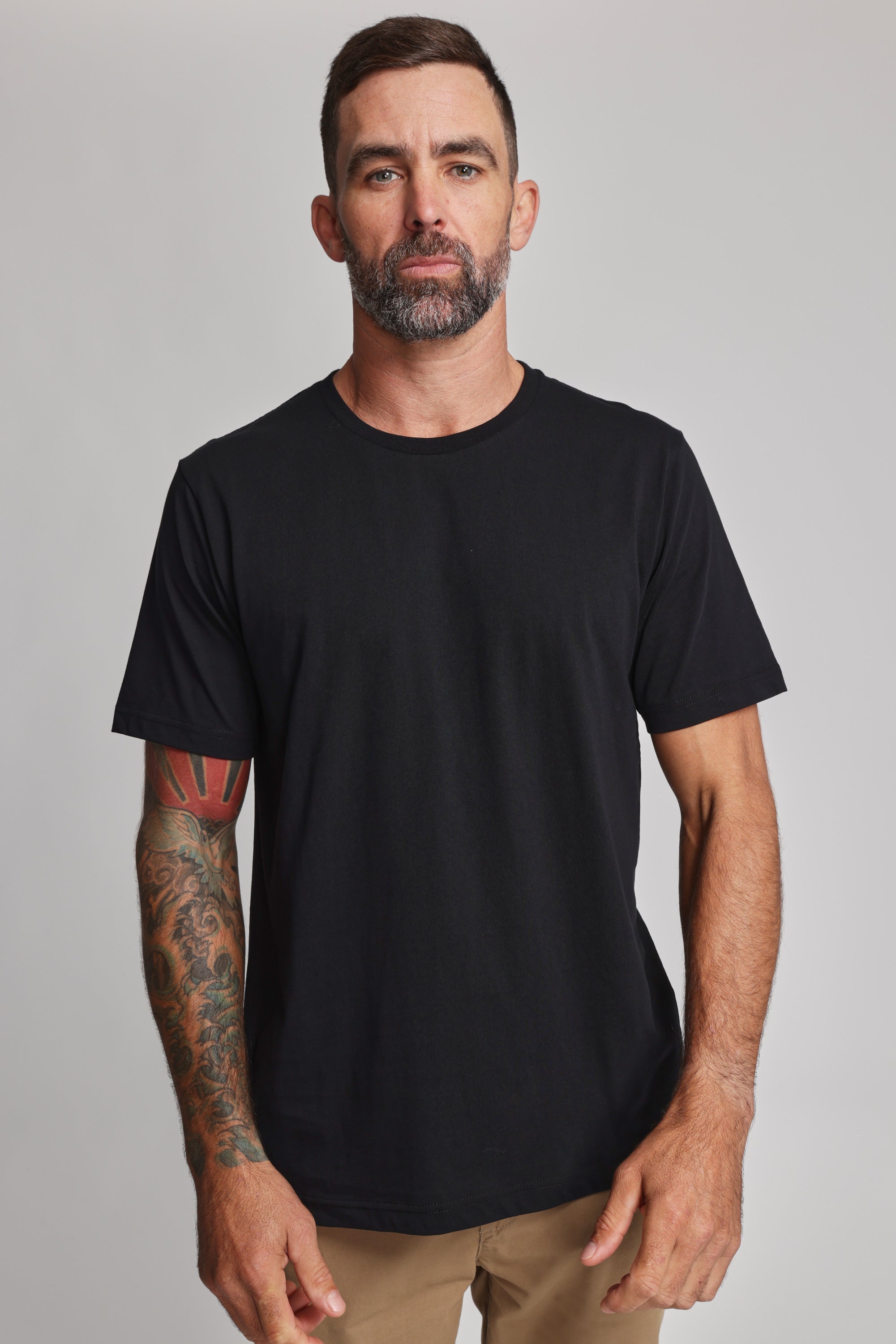 A man with short dark hair, a beard, and a tattooed right arm stands against a light gray background wearing the Barkers Organic Crew Tee and tan pants. He faces the camera with a neutral expression.