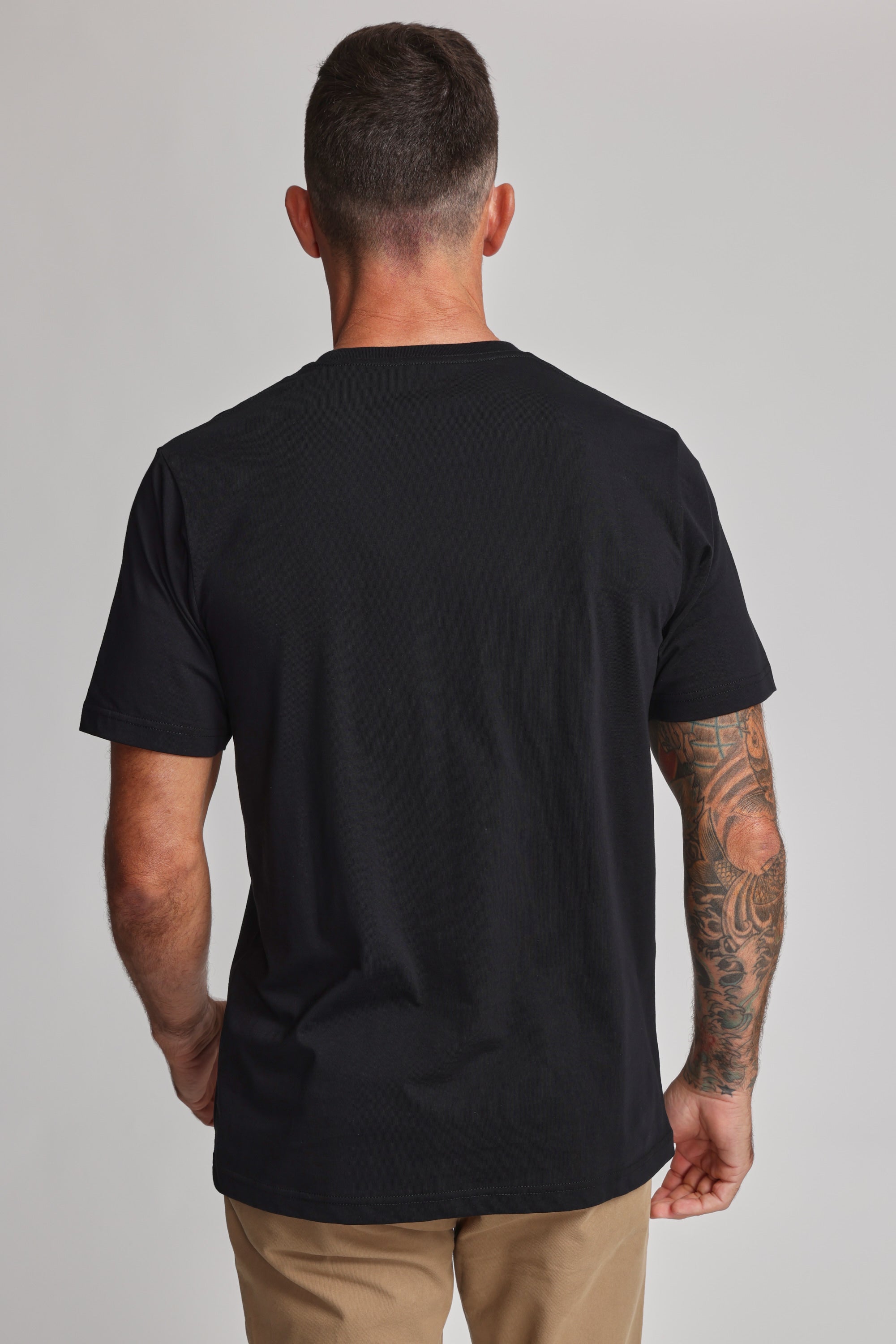 A man with short dark hair, a tattooed right arm, and tan pants stands with his back to the camera in a black Barkers Organic Crew Tee made from mid-weight organic cotton, set against a plain light background.
