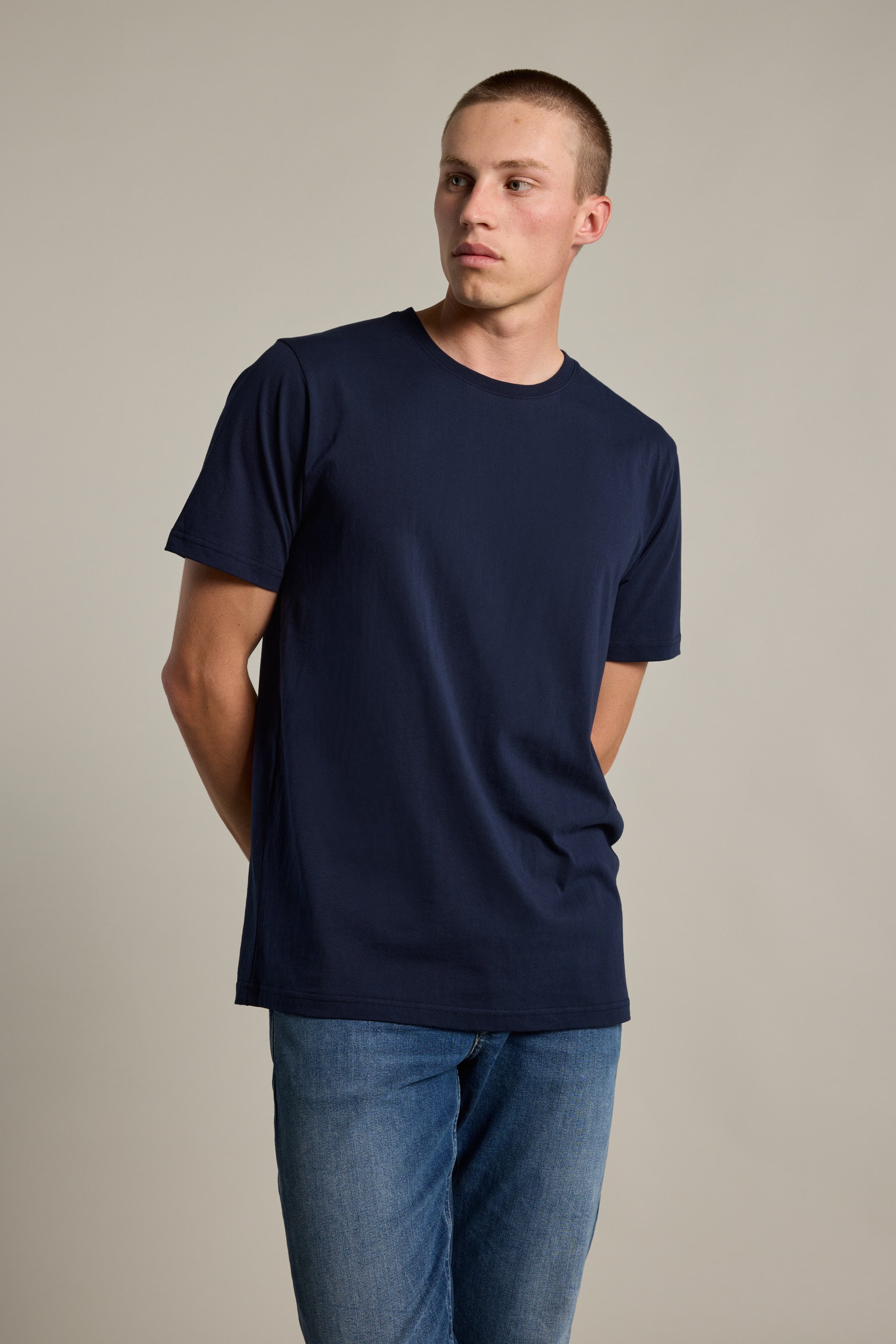 A man in a plain navy blue Barkers Organic Crew Tee and blue jeans stands against a light, neutral background with his hands behind his back, looking to the side.