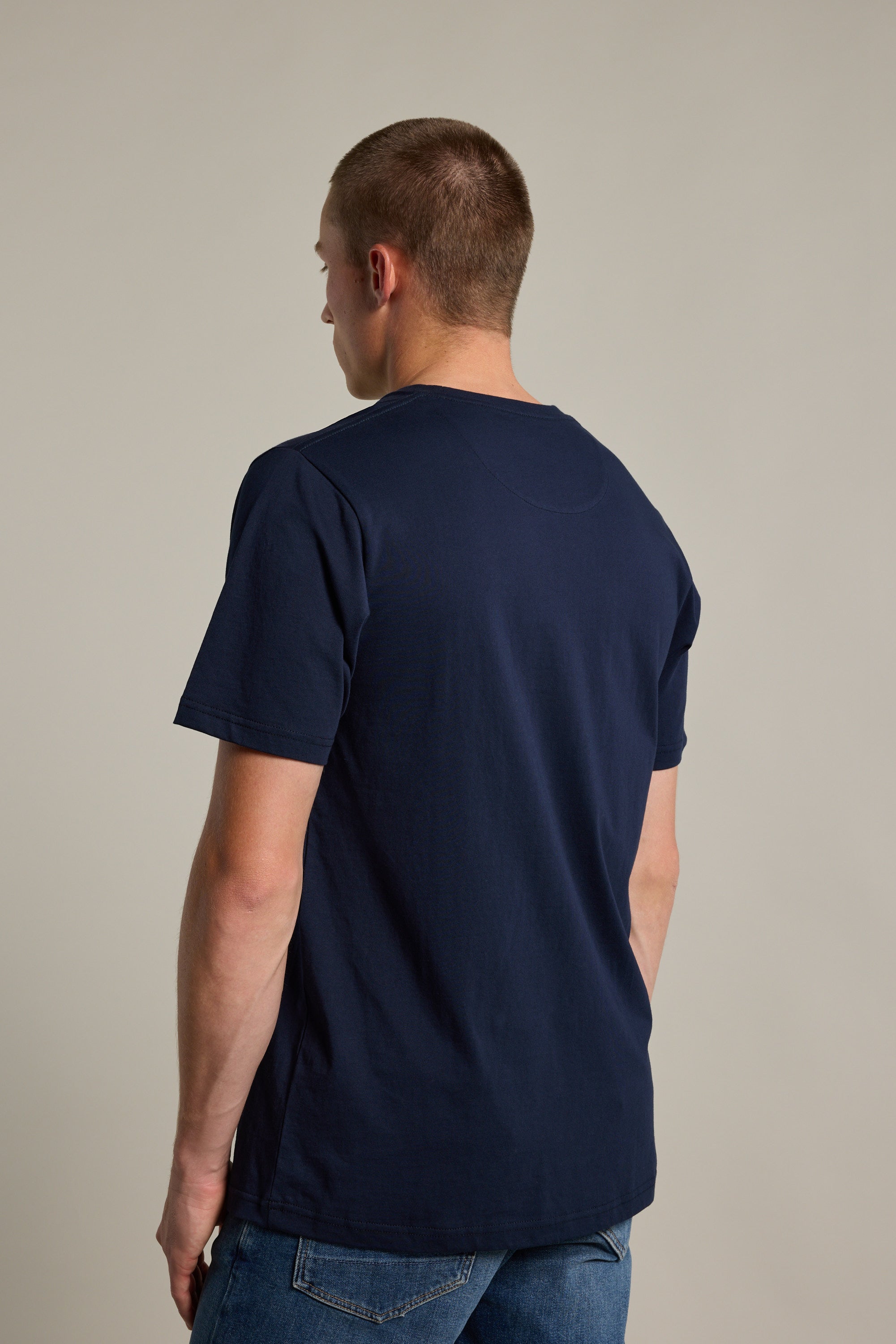 A person with short hair faces away, displaying the back of the Barkers Organic Crew Tee in navy blue with blue jeans against a plain, light background.